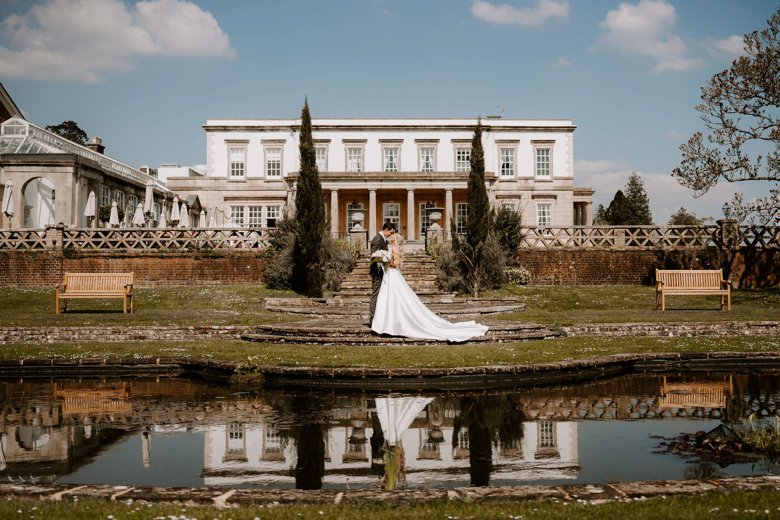 A bride and groom kiss on a flight of stone steps in front of the pale front of Buxted Park Hotel, with the house and their reflection in a long pond in the foreground