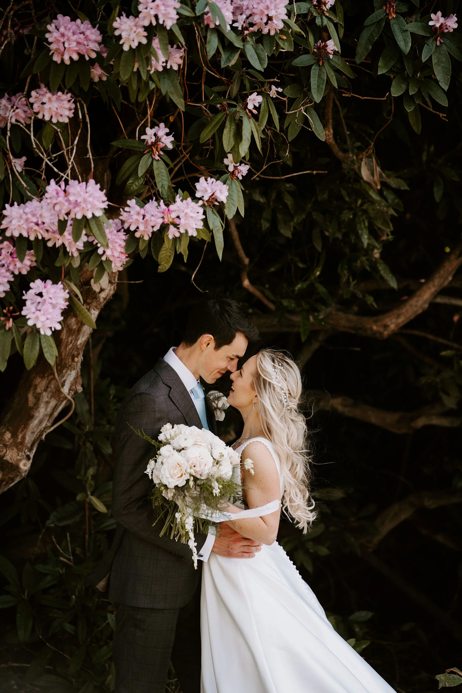 A groom in a grey check suit and a bride in a white gown holding a bouquet of cream roses stand nose to nose beneath a pink flowering rhododendron at Buxted Park Hotel near Uckfield