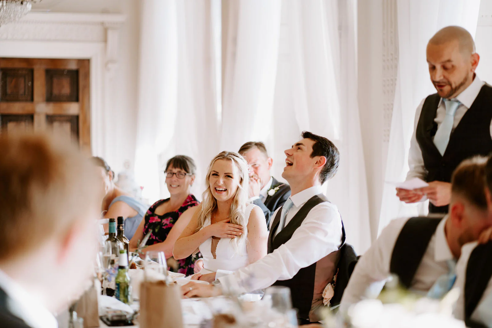 A bride laughs with a hand on her chest and a groom laughs beside her while a man in a waistcoat reads a speech at the top table at Buxted Park Hotel