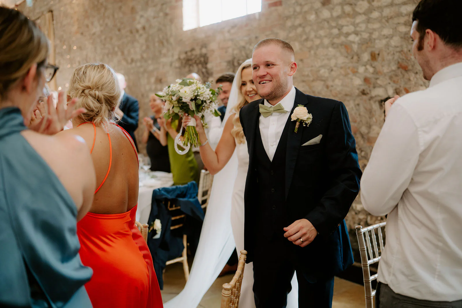 Laura holding a bouquet and Ricky in a black suit and green bow tie arrive smiling in the flint barn as friends and family gather around them