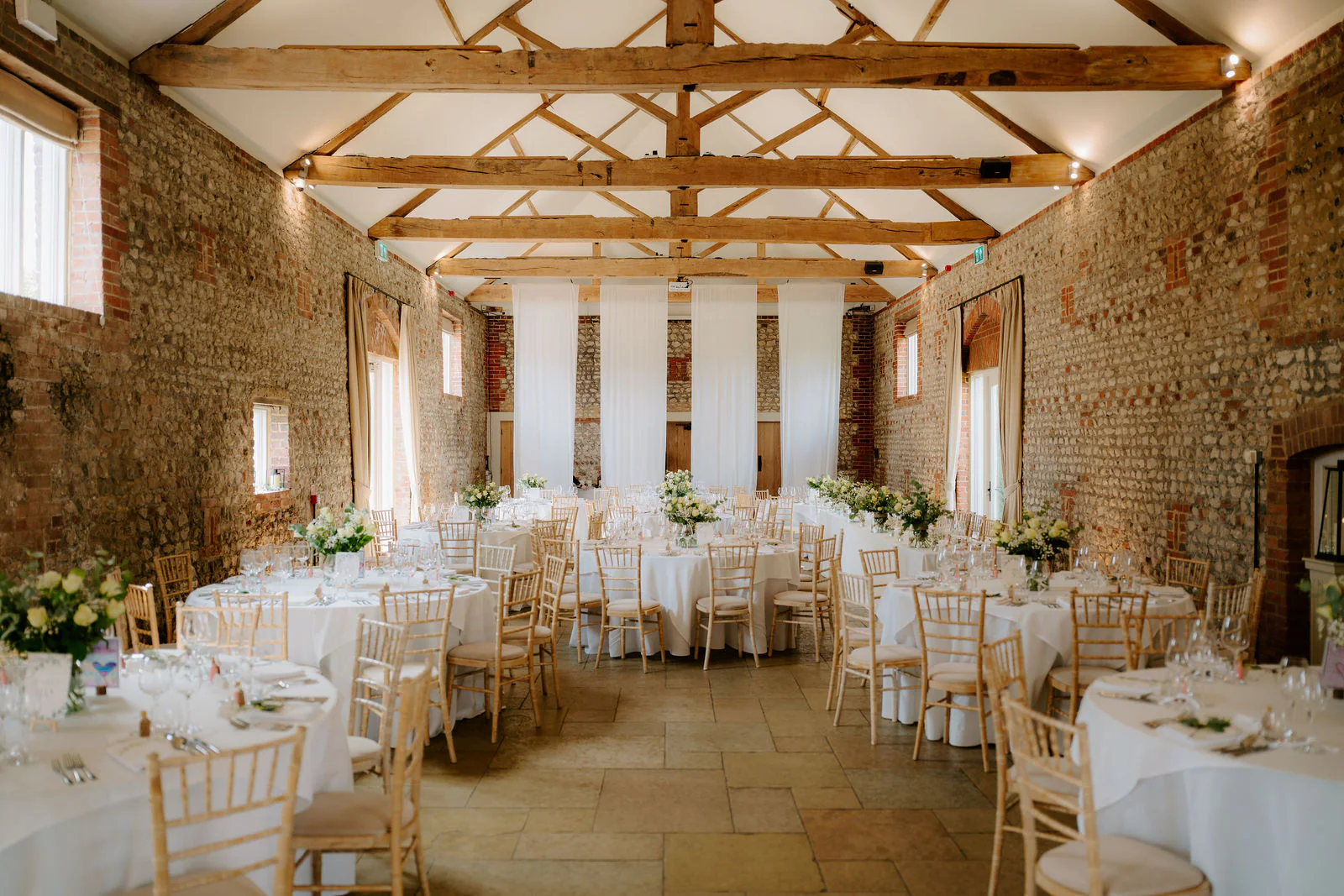 The long flint and brick barn at Farbridge laid with round tables, white chairs and flowers beneath exposed oak trusses