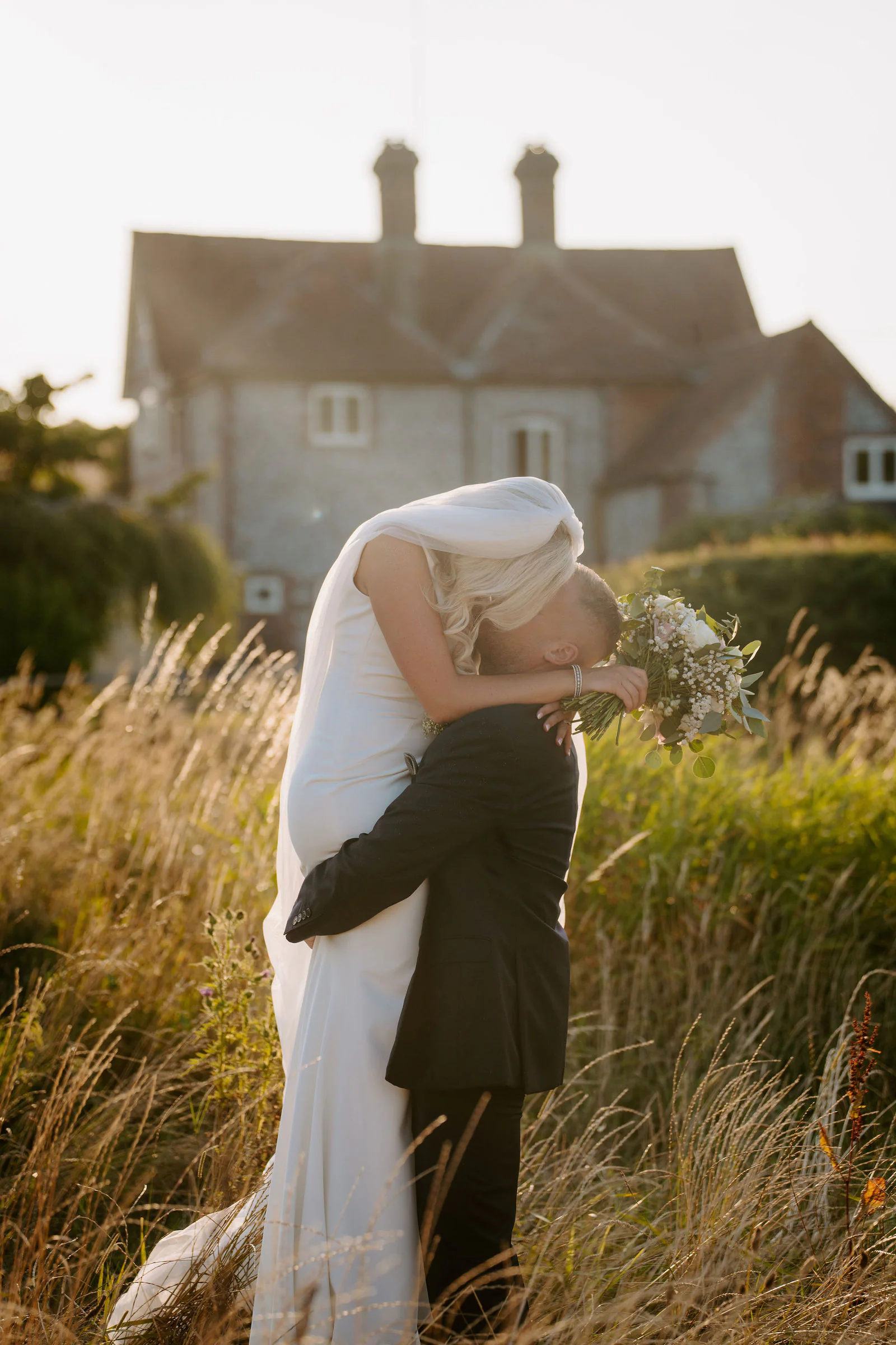 Ricky lifts Laura into a kiss in tall grass in front of a stone farmhouse with two chimneys, her veil swept across her face