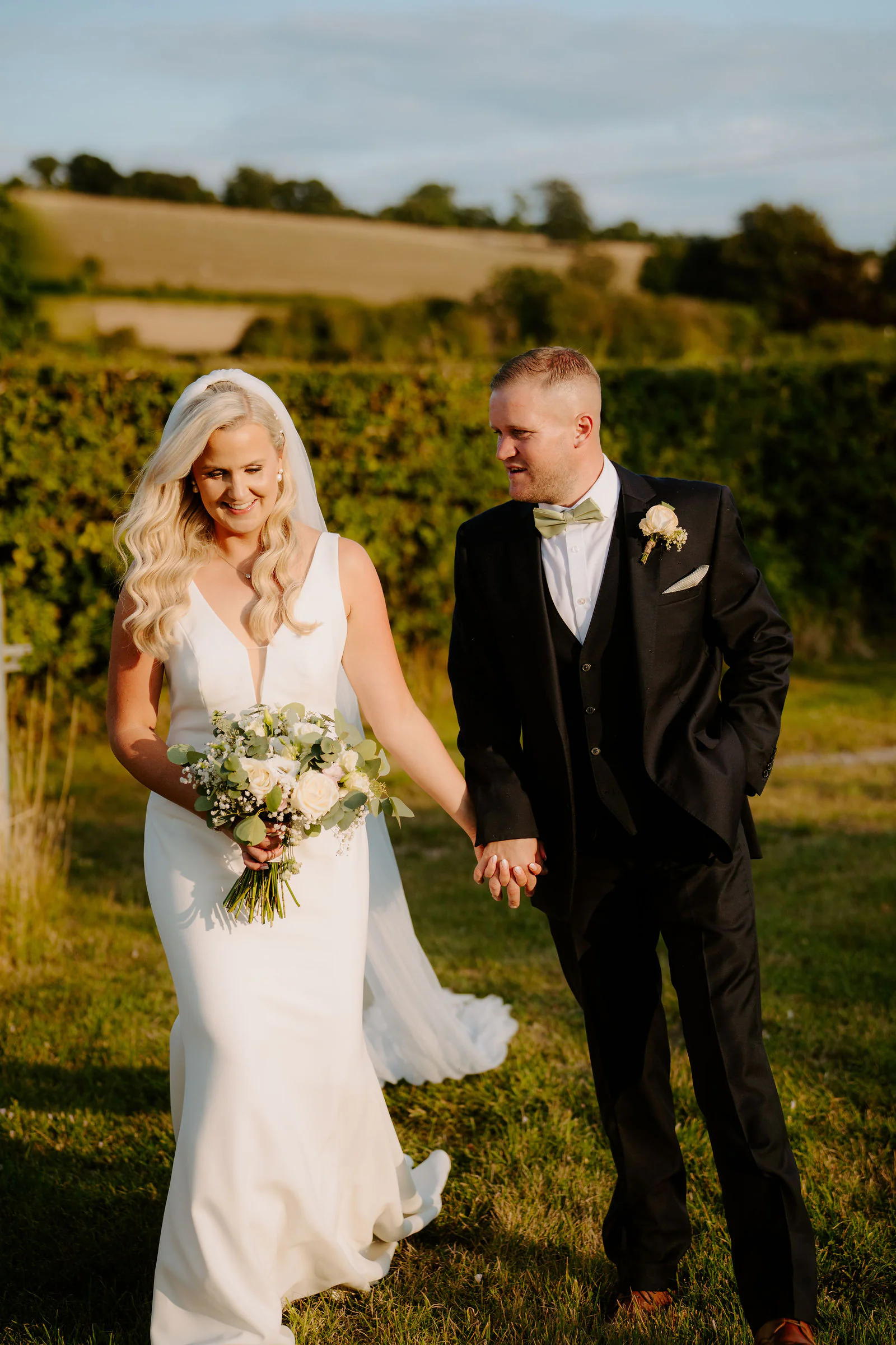 Laura and Ricky walk hand in hand across a sunlit field beside a hedge with rolling downland behind them