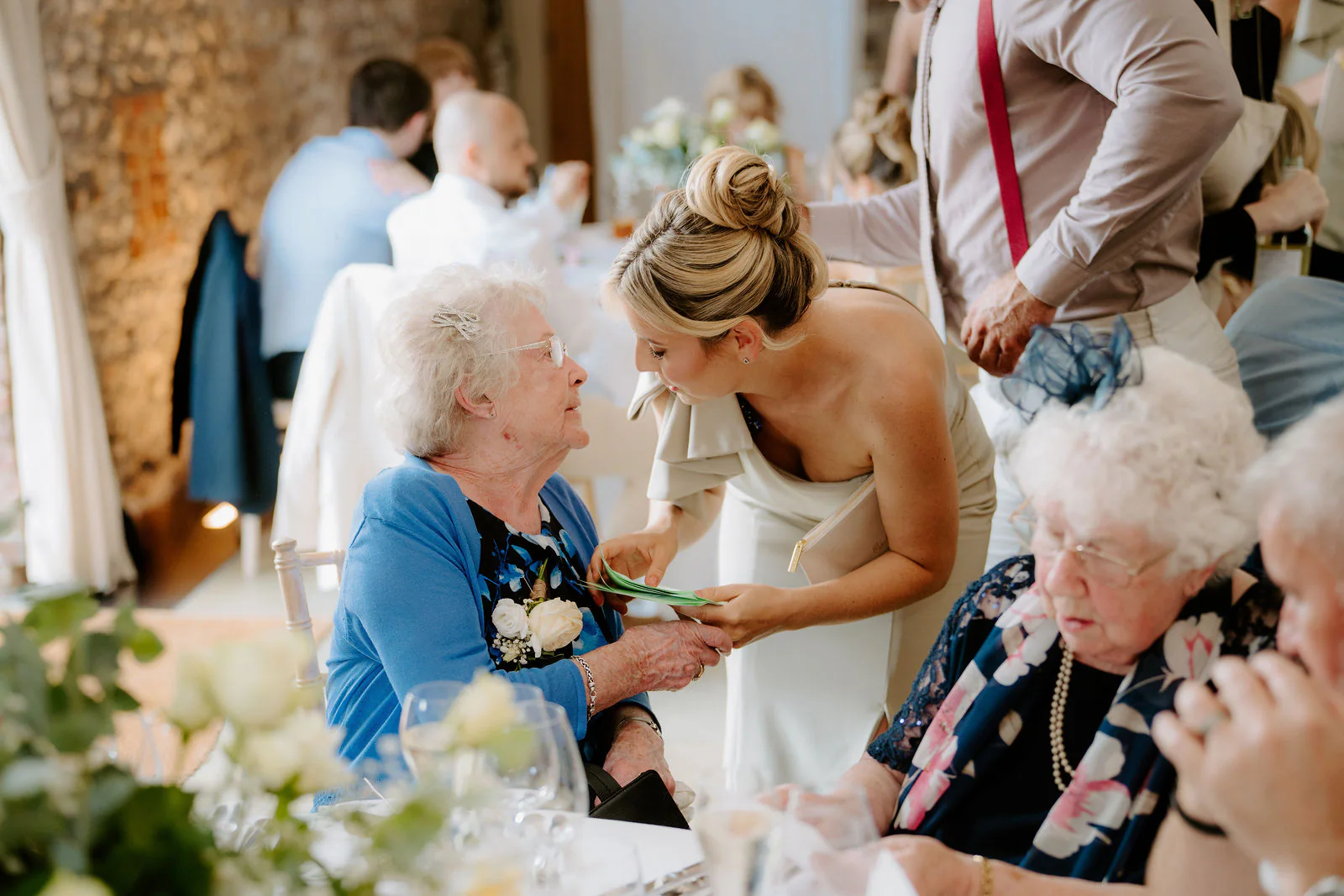A bridesmaid in a pale dress leans down to talk to two grandmothers seated at a table in the barn at Farbridge