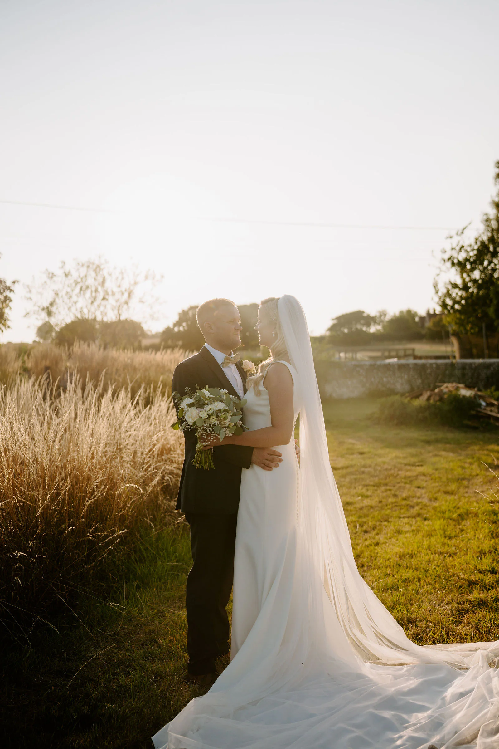 Laura and Ricky embrace in tall golden grasses with a stone wall and fields behind them at sunset