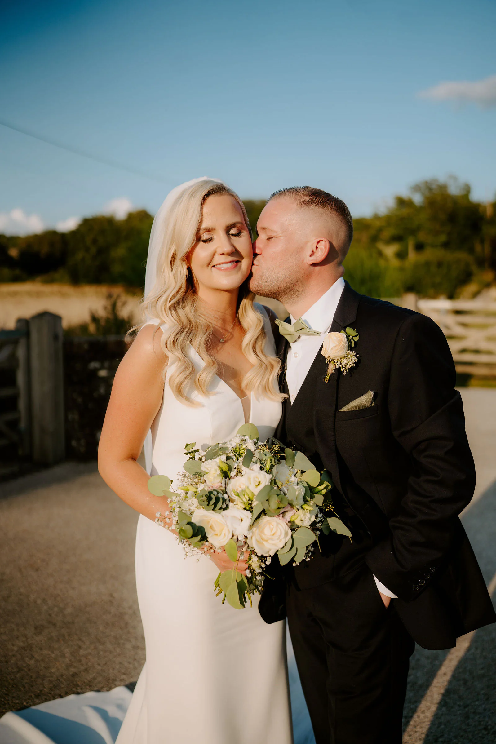 Ricky kisses Laura's cheek as she smiles with a bouquet of white and green flowers on a gravel drive at Farbridge