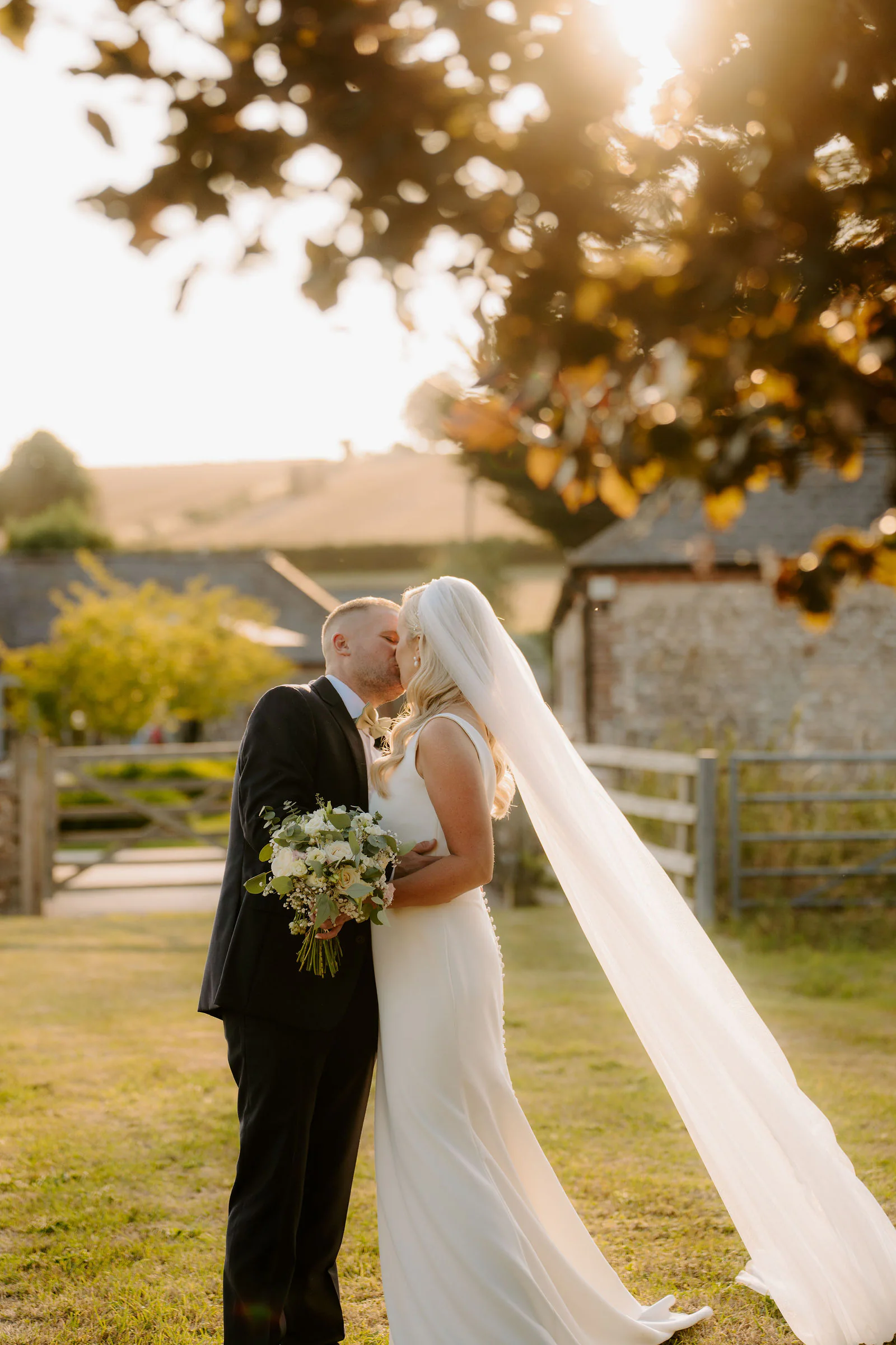 Laura and Ricky kiss in the low evening sun in front of a flint farm building and a wooden gate at Farbridge, her long veil lifted by the breeze