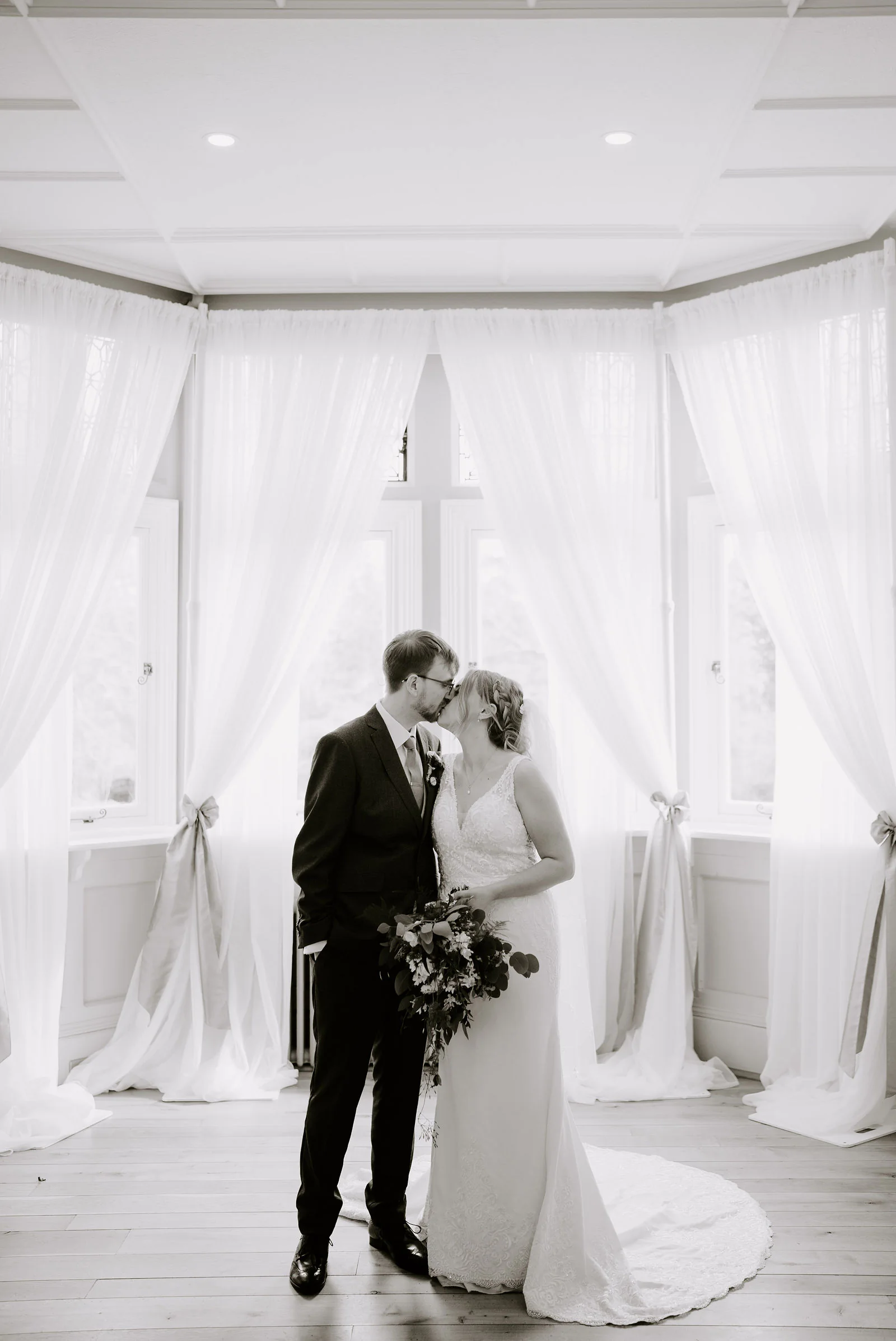 A black and white photograph of a bride and groom kissing in front of a bay window hung with white curtains tied with bows, her train spread on a wooden floor, at The Ravenswood