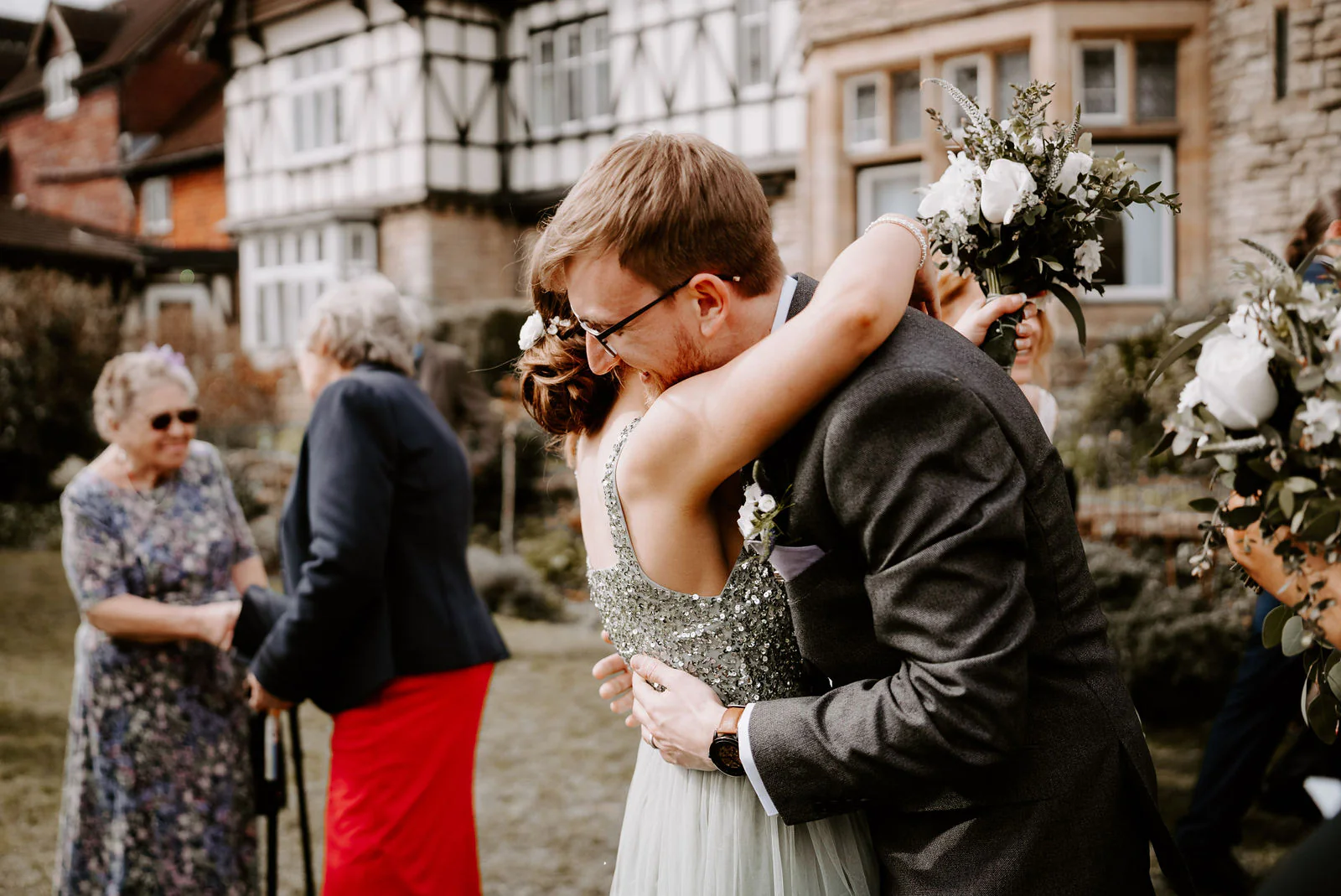 A groom is hugged by a bridesmaid in a sequin dress on the lawn in front of a half-timbered and stone house at The Ravenswood while guests chat behind them