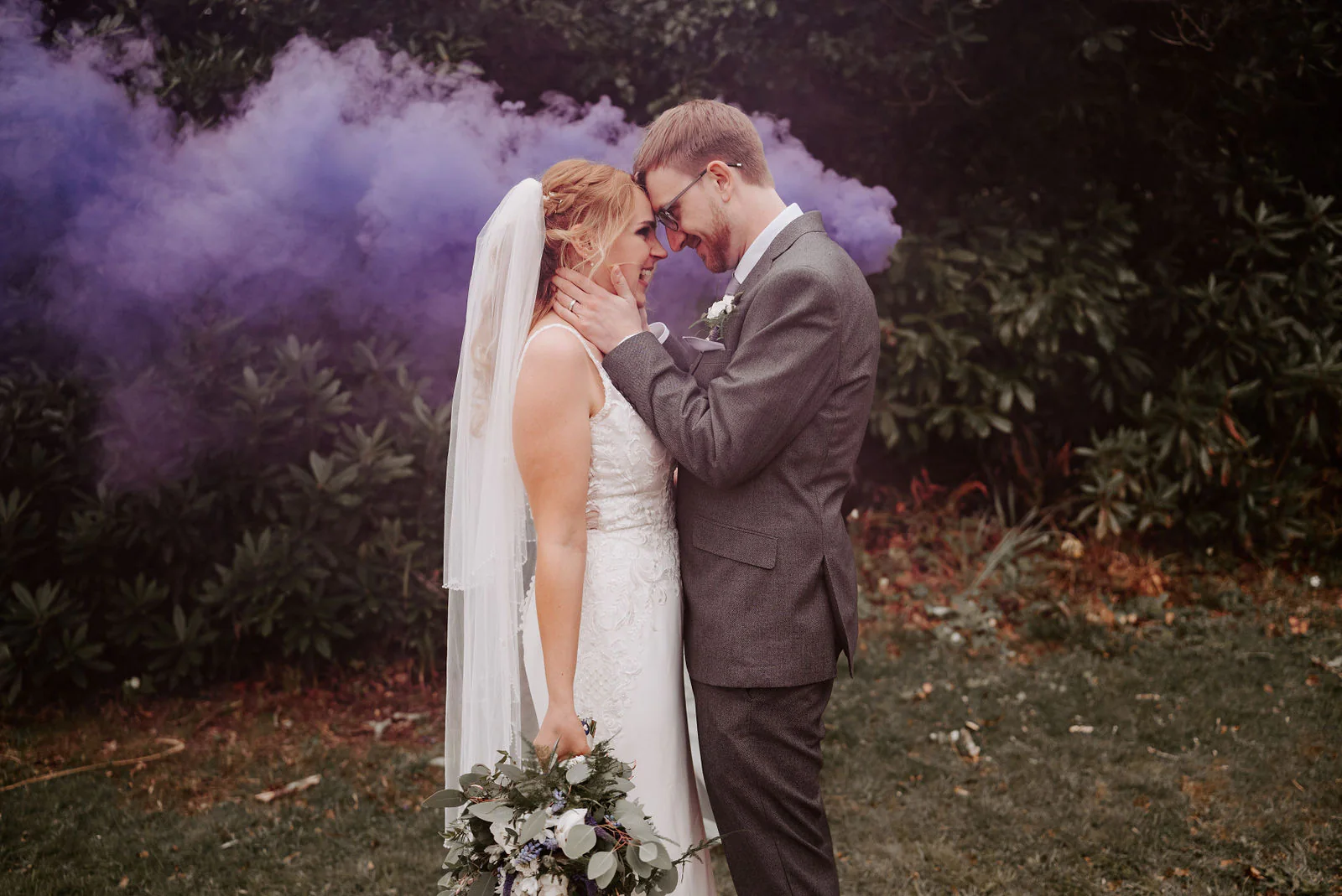 A groom in a grey suit and glasses touches foreheads with a bride in a lace gown and veil in front of dark shrubs and a cloud of purple smoke in the grounds of The Ravenswood near East Grinstead
