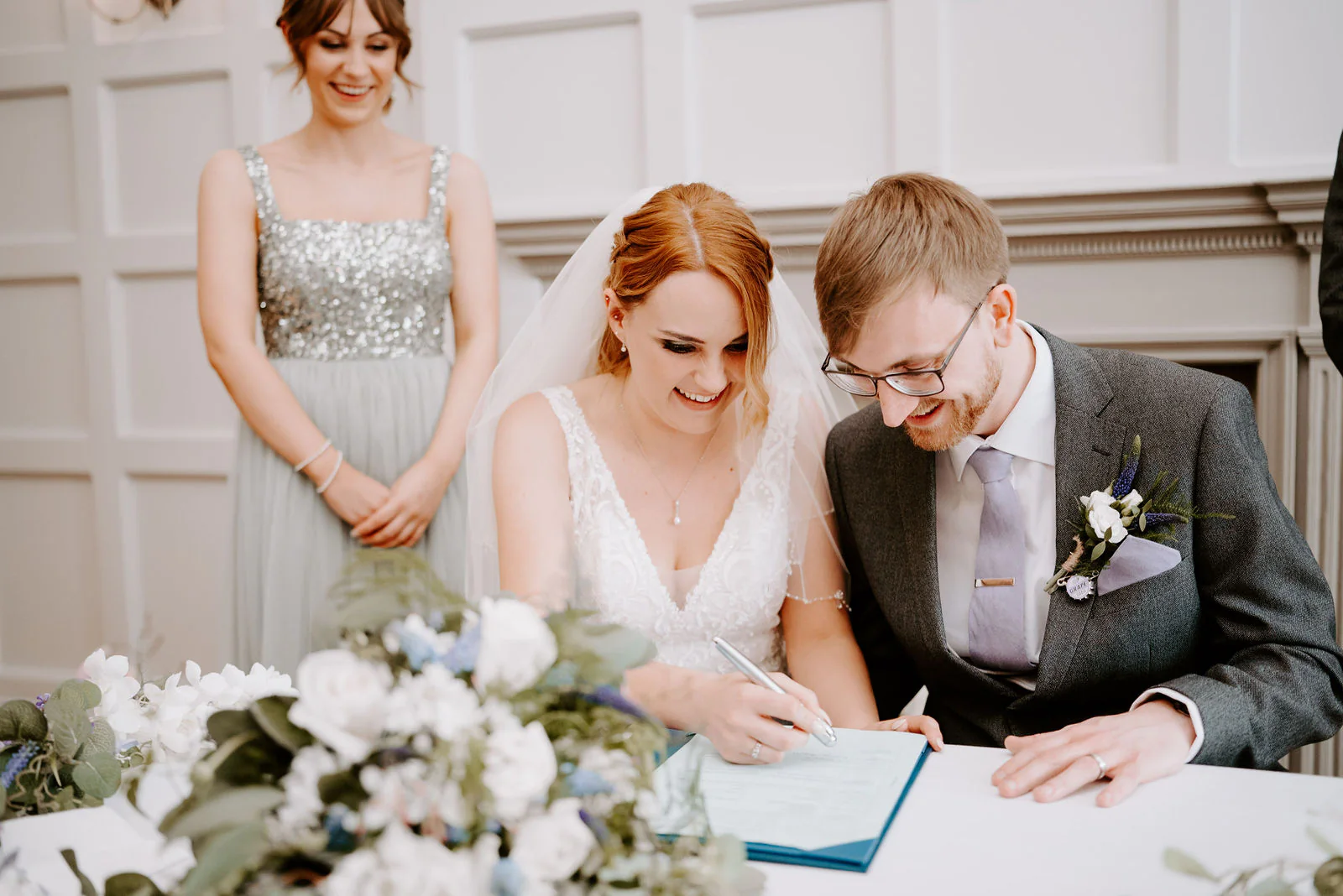 A bride in a lace gown and a groom in glasses and a grey suit laugh as they sign the register while a bridesmaid in a sequin dress looks on at The Ravenswood