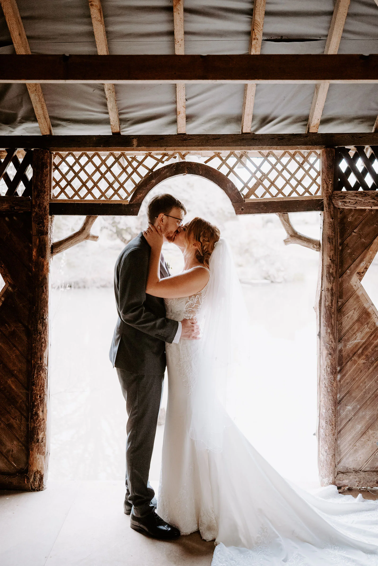 A bride and groom kiss in the timber and lattice doorway of a wooden lakeside summerhouse at The Ravenswood, with bright water behind them