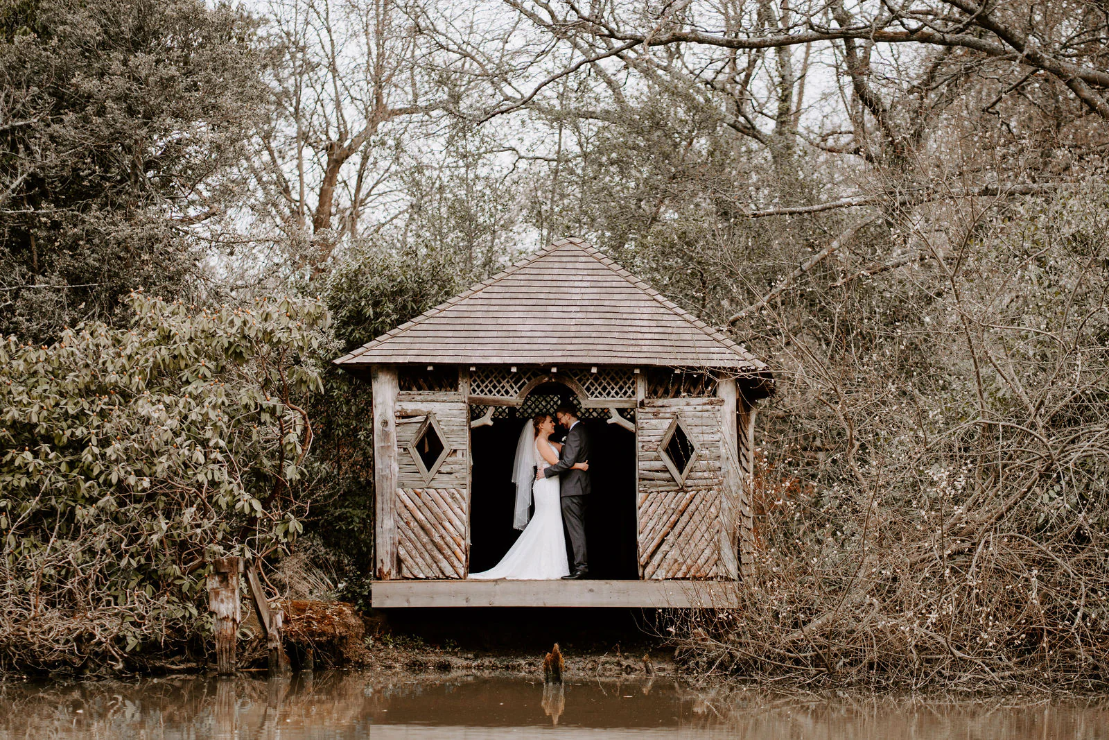 A bride and groom embrace inside a small wooden summerhouse seen from across the water, among bare branches and rhododendron, in the grounds of The Ravenswood