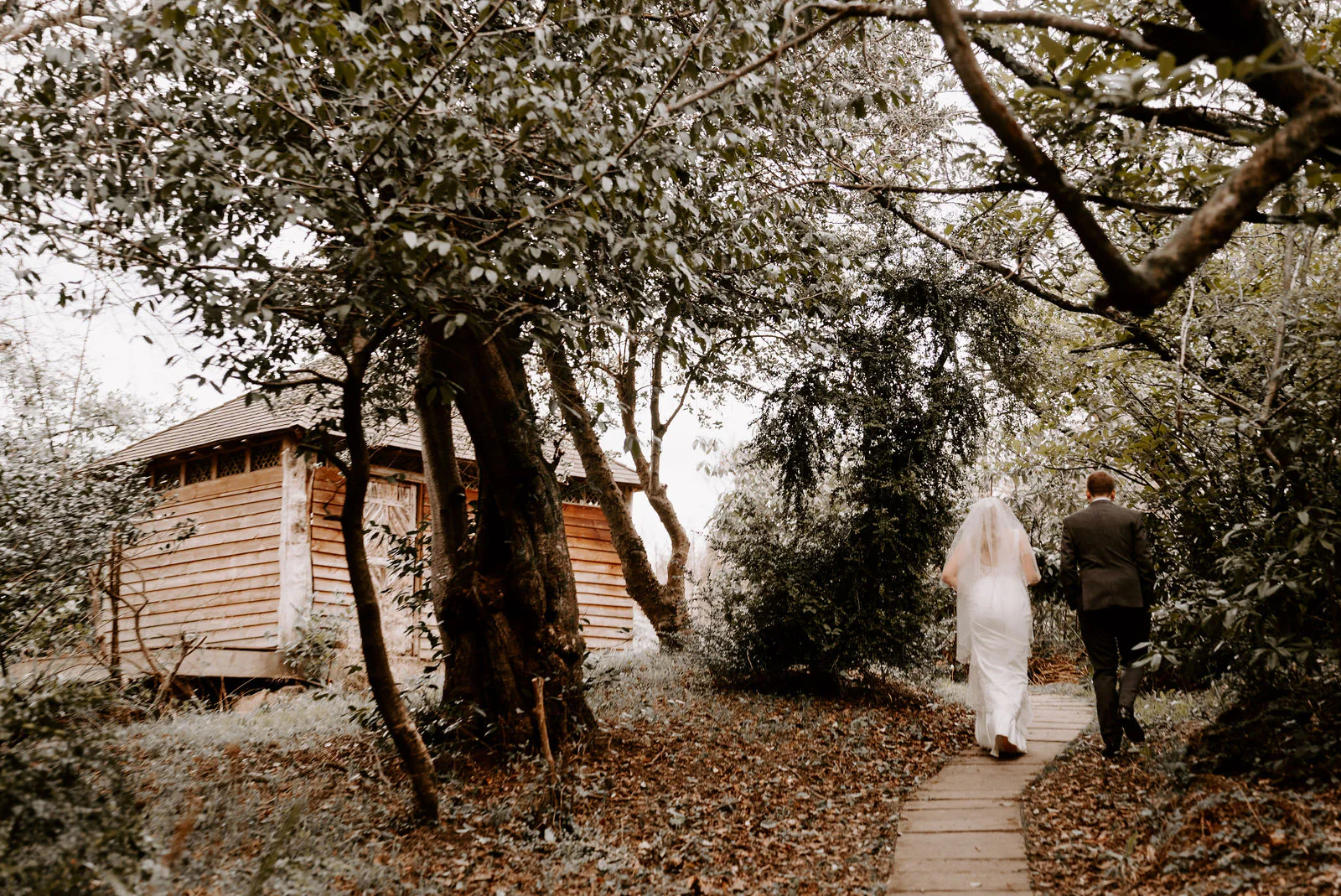 A bride in a veil and a groom in a dark suit walk away from the camera along a flagstone path under old trees, past a wooden summerhouse, in the grounds of The Ravenswood