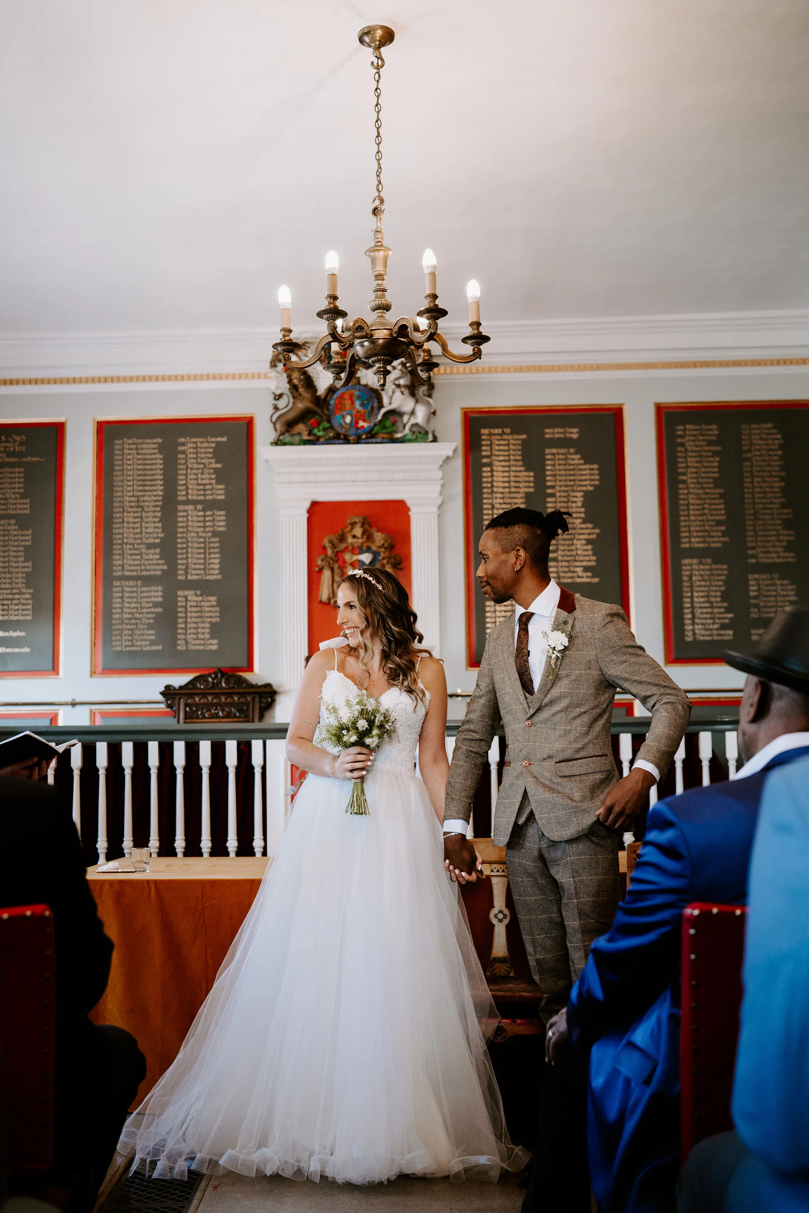Alexandra and Row stand hand in hand in a panelled room with a chandelier and boards of names on the walls, guests seated beside them