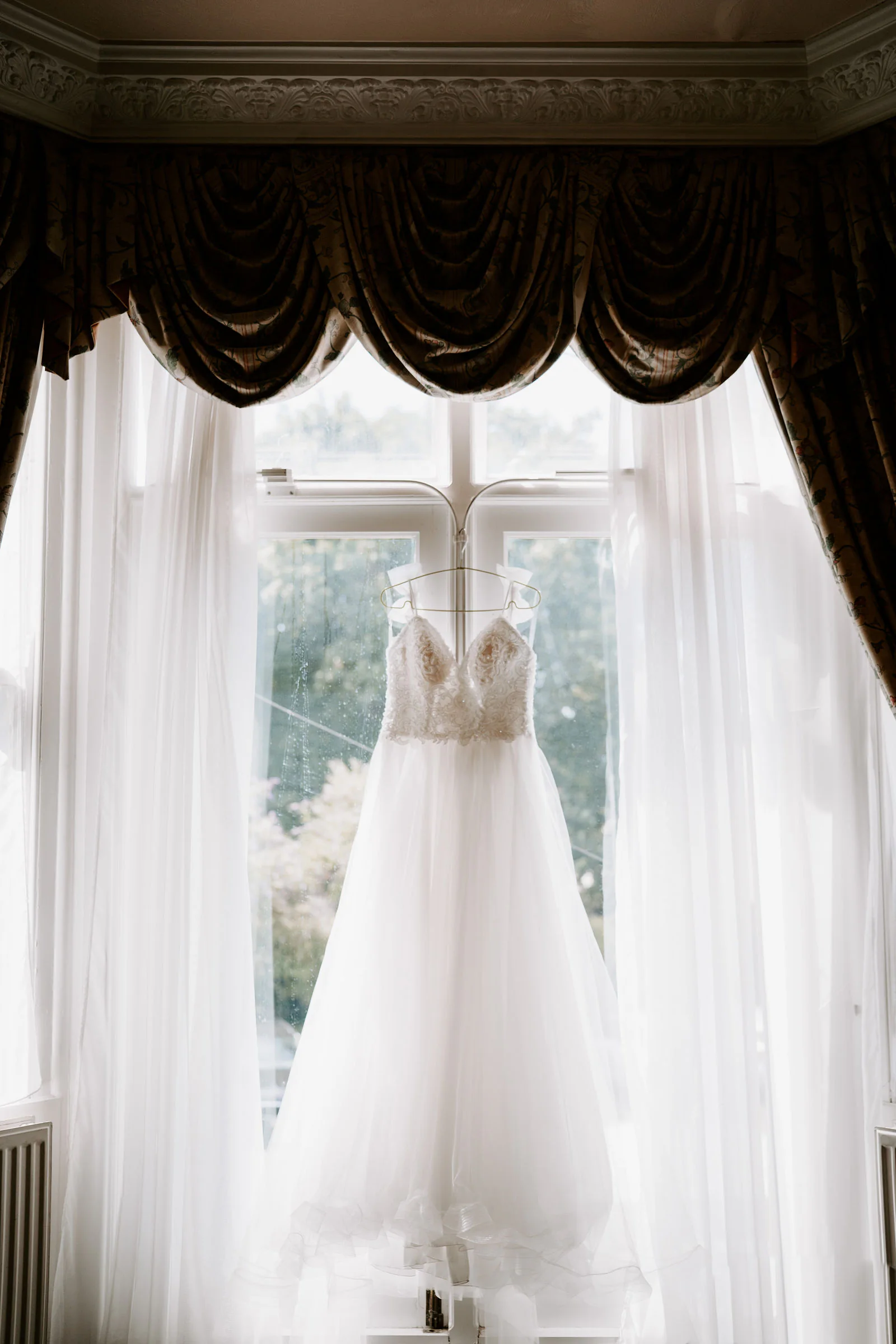 A wedding dress hangs in a tall bay window with dark swagged curtains and white voiles before the wedding in Rye