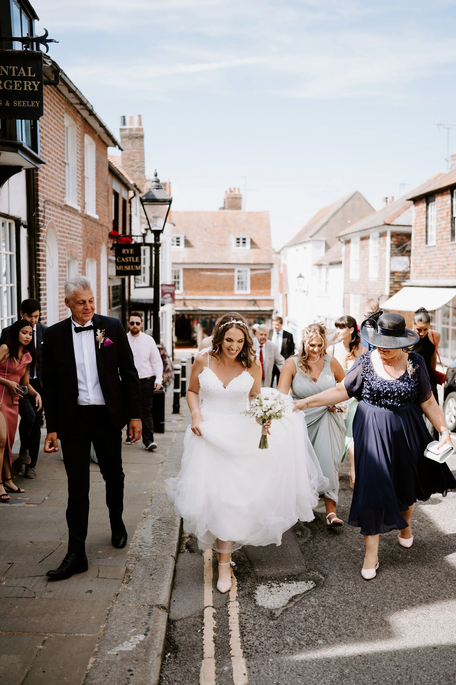 Alexandra in her wedding dress lifts her skirt as she walks down a Rye street with her father, bridesmaids and family beside her