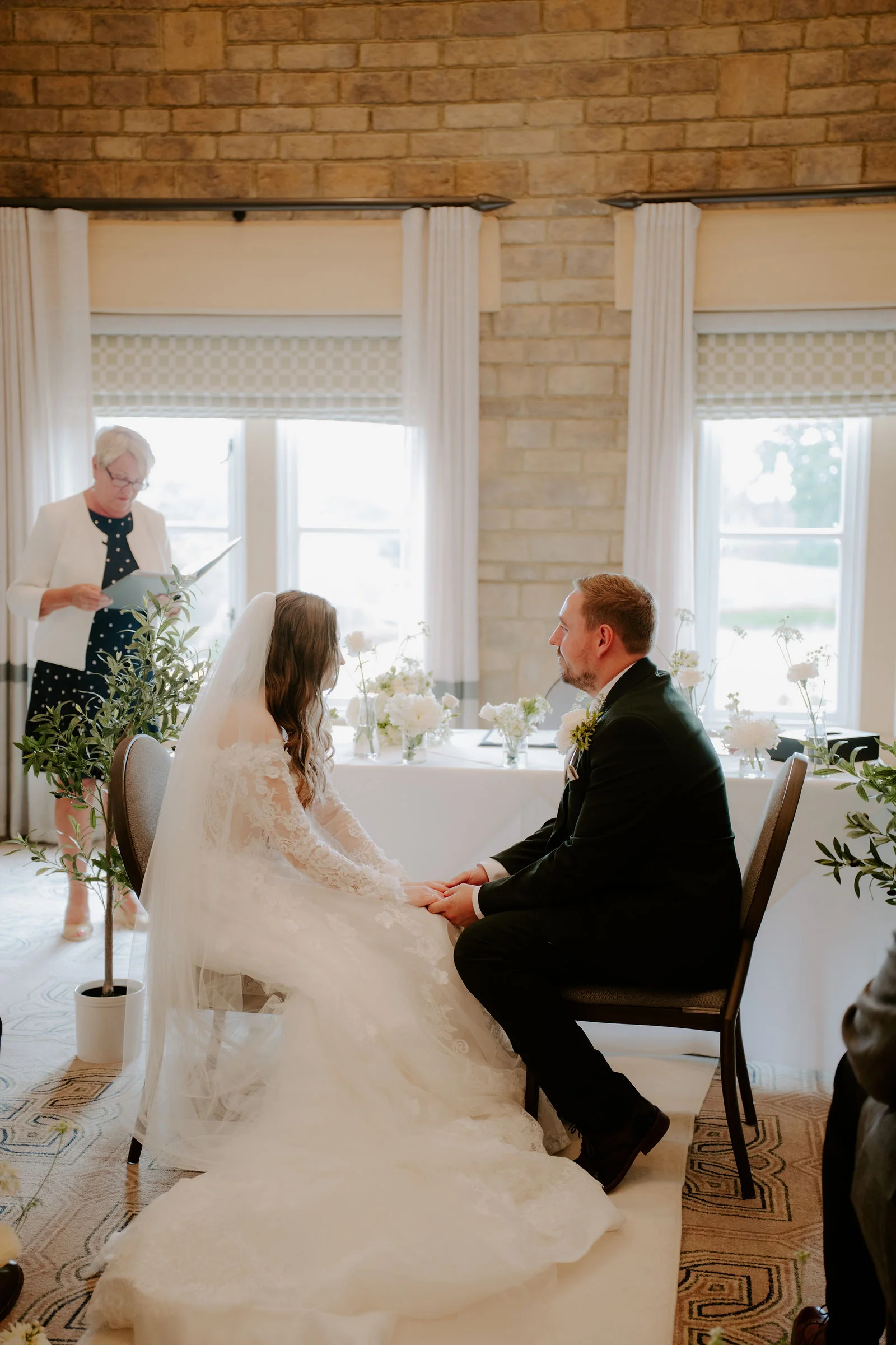 A bride in a veil and a long lace train and a groom sit holding hands at a table in front of tall windows in a stone-walled room while a woman in a white jacket reads to them