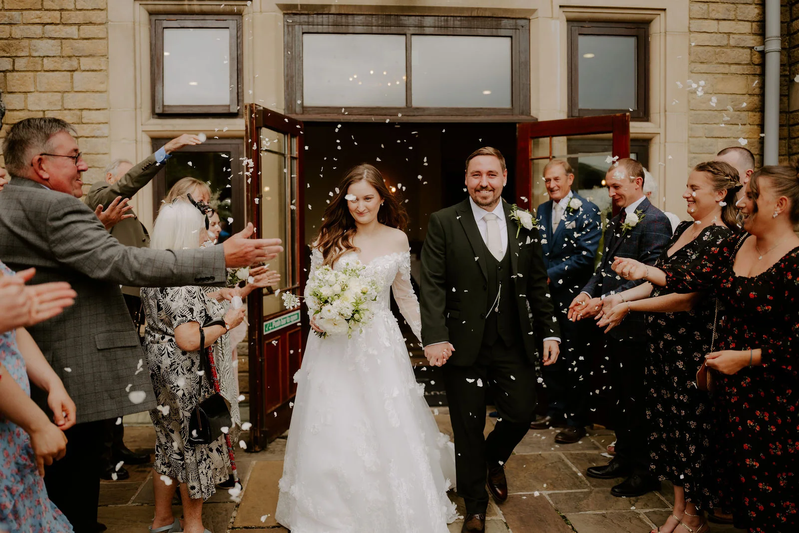 A bride in an off-the-shoulder lace gown holding a bouquet of white flowers and a groom in a dark green suit walk out through open double doors of a stone house in a shower of confetti, with guests lining both sides