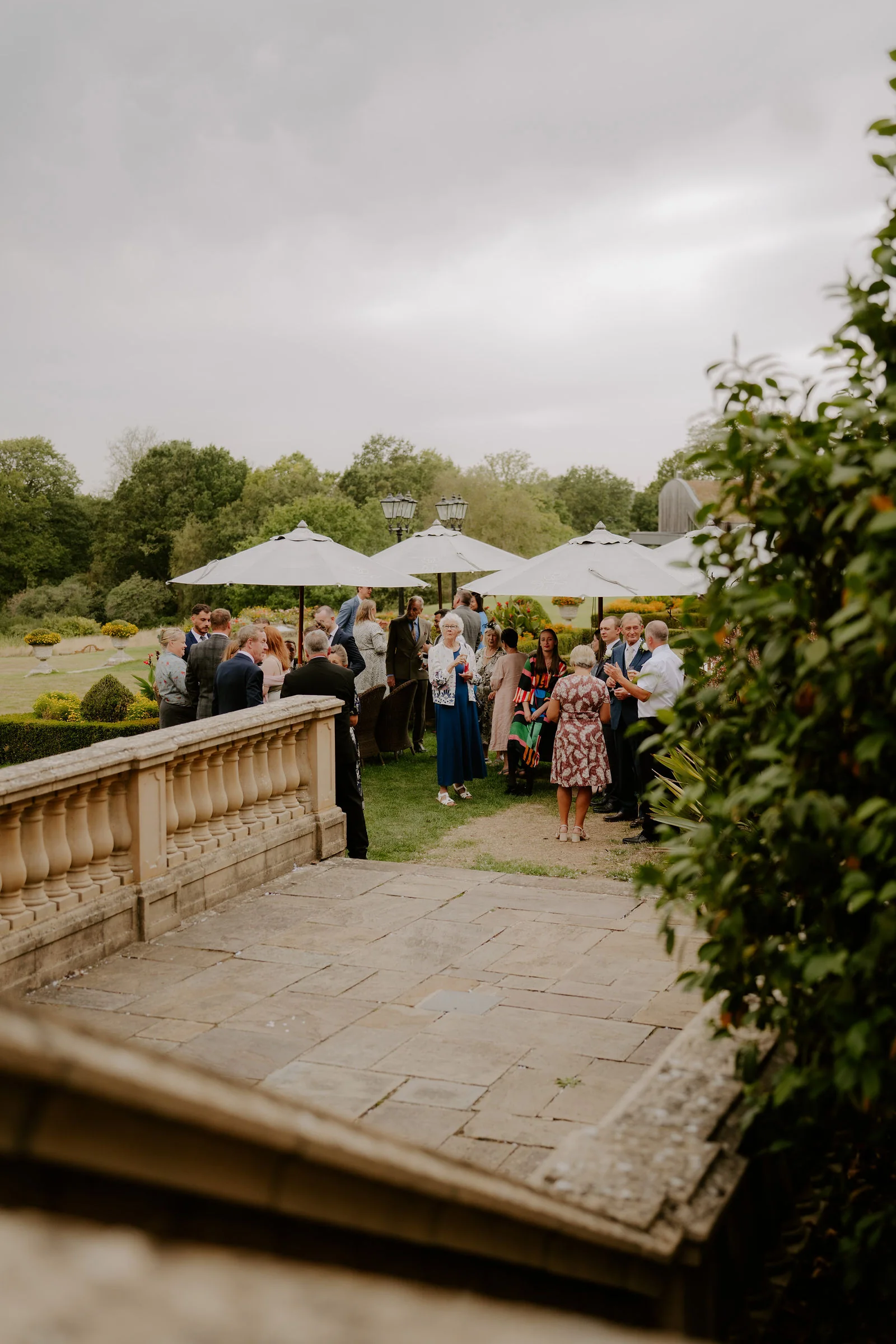 Wedding guests gather on a stone terrace beside a balustrade under white parasols, with a terraced garden and trees below under an overcast sky