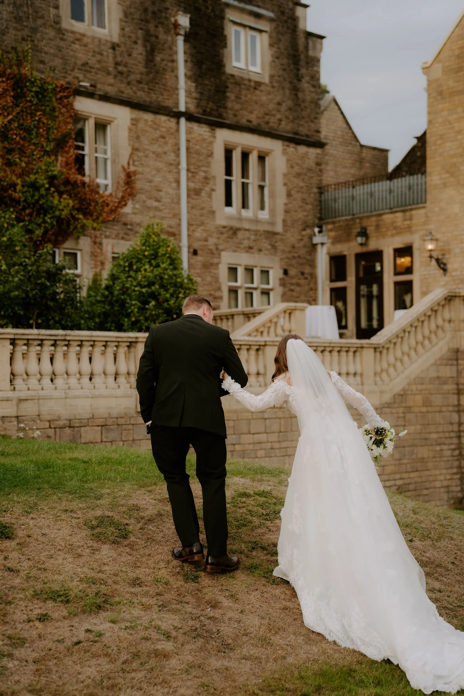 Seen from behind, a bride in a long veil and a groom in a dark suit walk up a grassy bank beside a stone balustrade, with the stone house above them