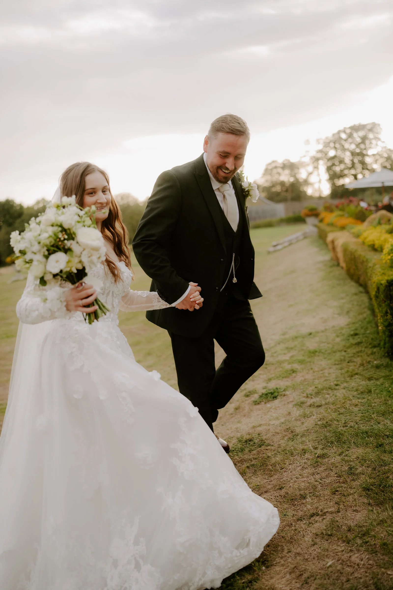 A smiling bride holding a bouquet of white flowers and her skirt walks with a groom in a dark suit on grass in soft light, with colourful flower beds behind them
