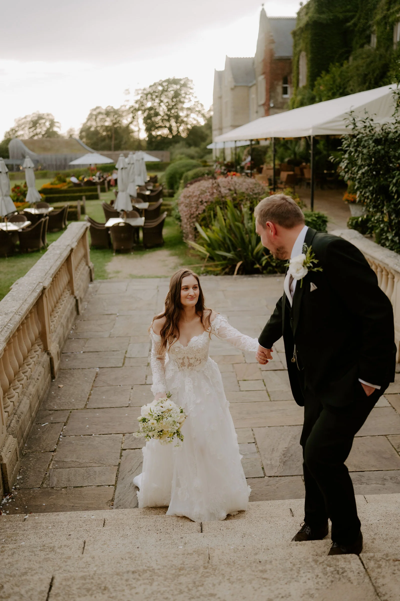 A groom takes the hand of his bride in a lace gown as she climbs wide stone steps beside a balustrade towards a terrace with white parasols and an ivy-covered stone house