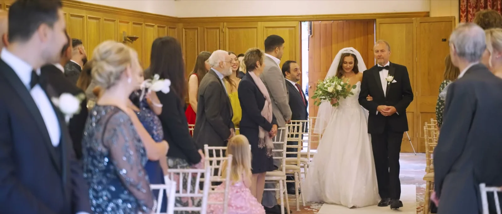A bride carrying white flowers walks down the aisle on the arm of a man in a dark suit in a wood-panelled room, guests standing on both sides