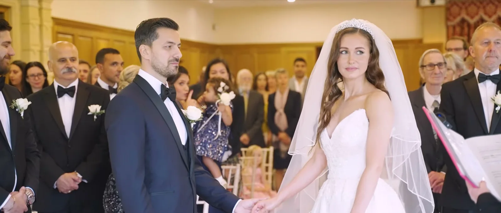 A groom and a bride in a veil and tiara hold hands at the front of the ceremony room while their guests watch