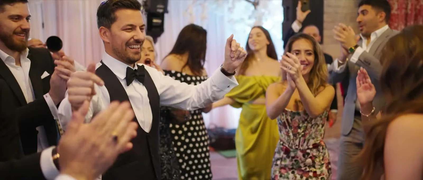 A groom in a waistcoat and bow tie dances with his hand raised as guests clap around him