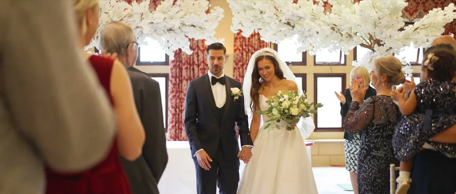 A bride holding a bouquet and a groom walk hand in hand past clapping guests beneath white blossom branches