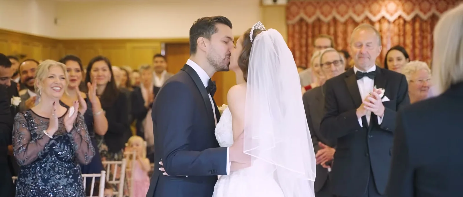 A couple kiss in the ceremony room while guests behind them applaud