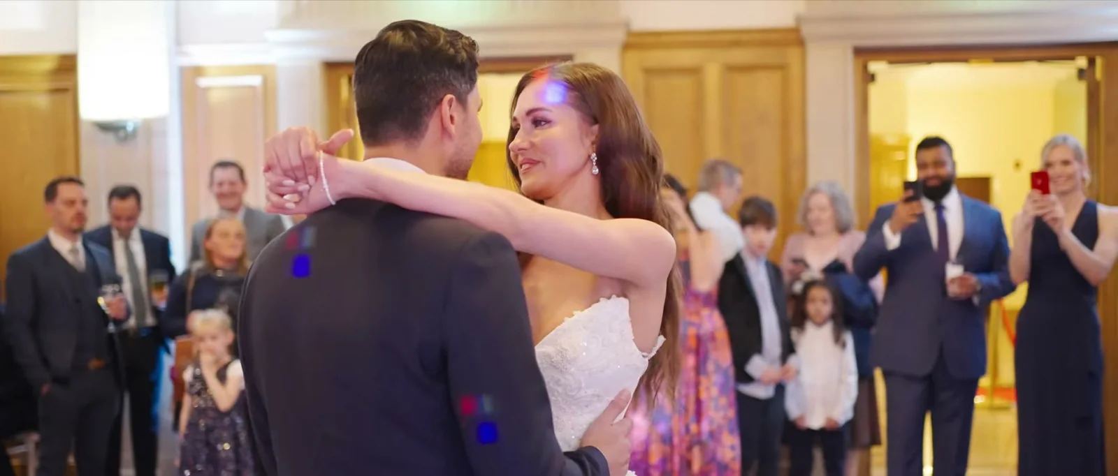 A groom and a bride share their first dance in a panelled room while guests stand in a ring around them