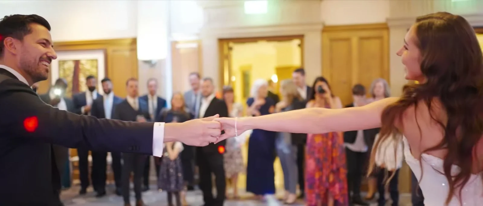 A groom and a bride reach for each other's hands at arm's length on the dance floor as guests watch