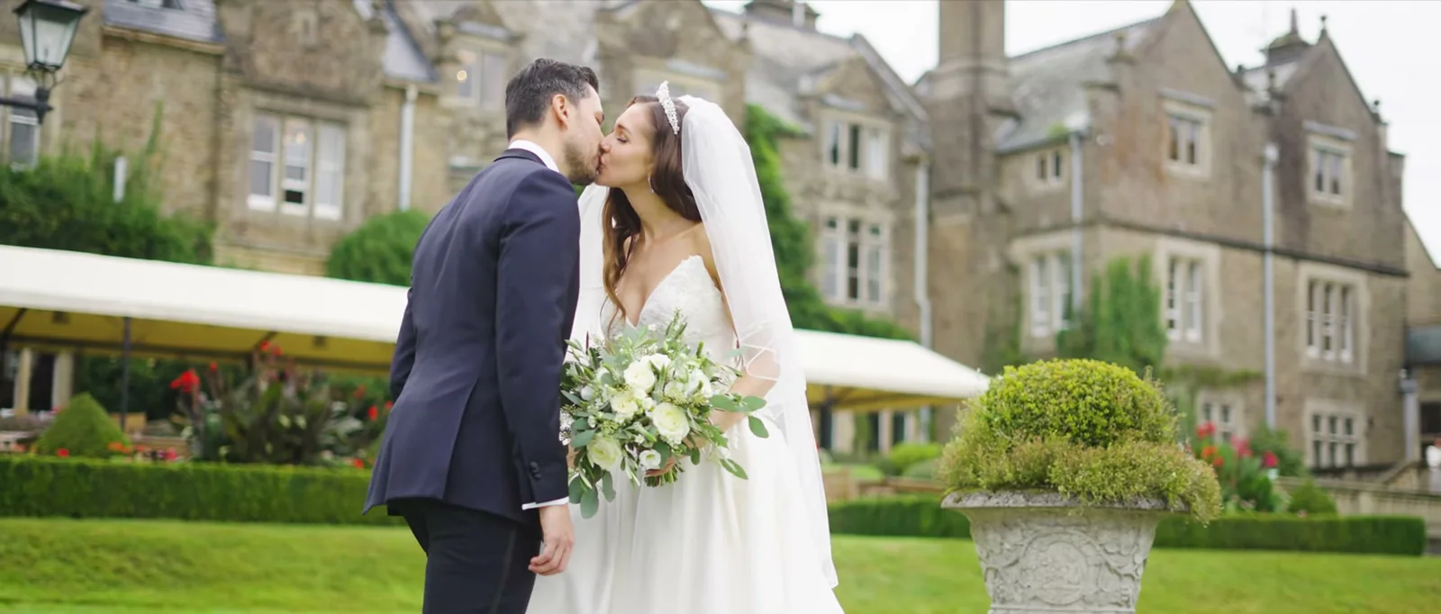 A couple kiss on the lawn with a bouquet of white flowers between them and the stone house behind