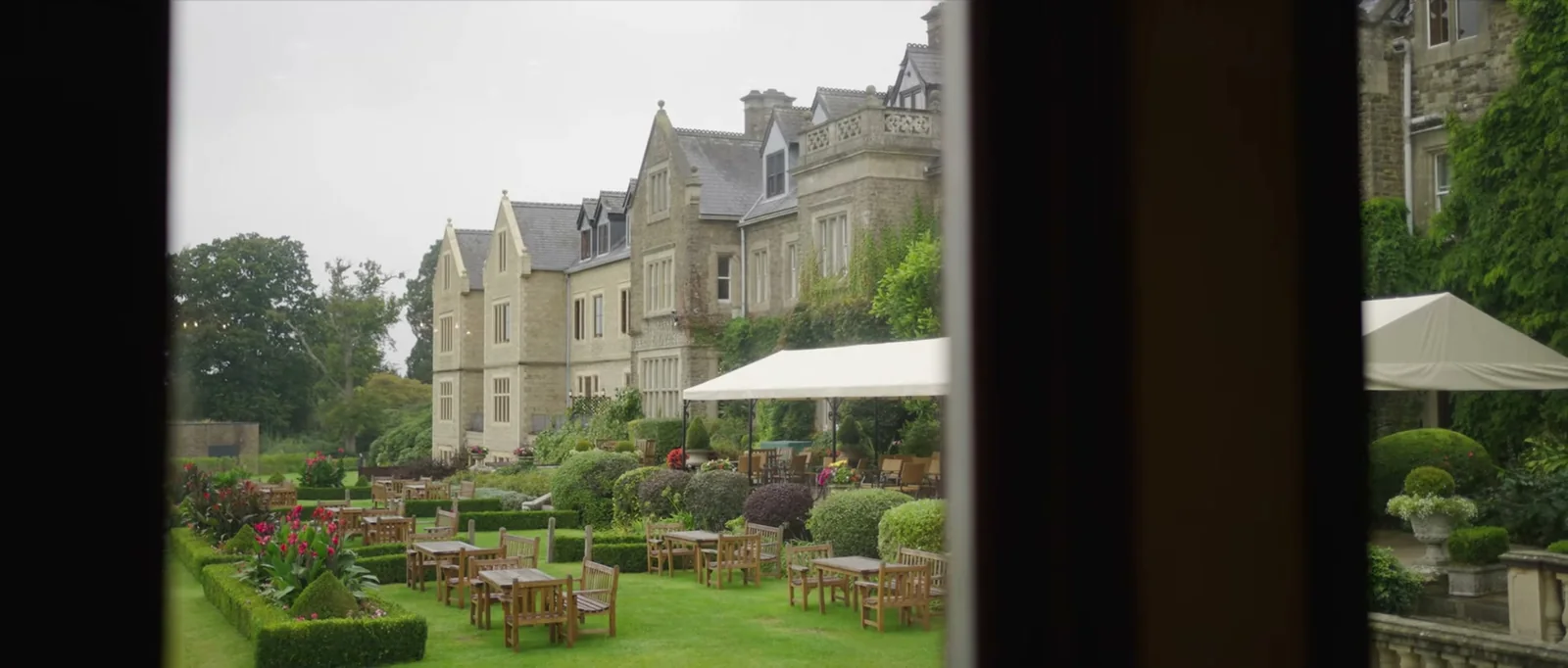 The stone front of South Lodge seen through a window, with a terraced garden, outdoor tables and a white canopy below