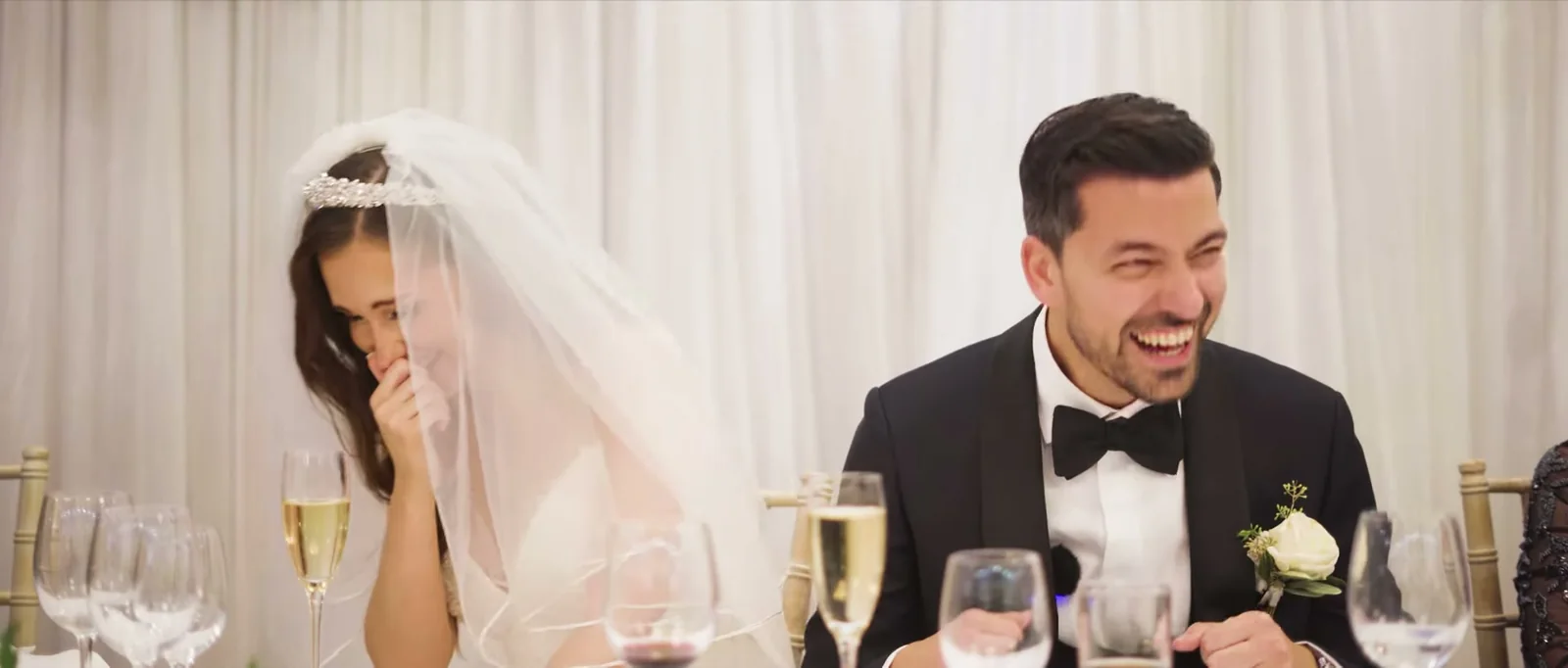 A bride covers her face as her groom laughs beside her at the top table in front of a white drape
