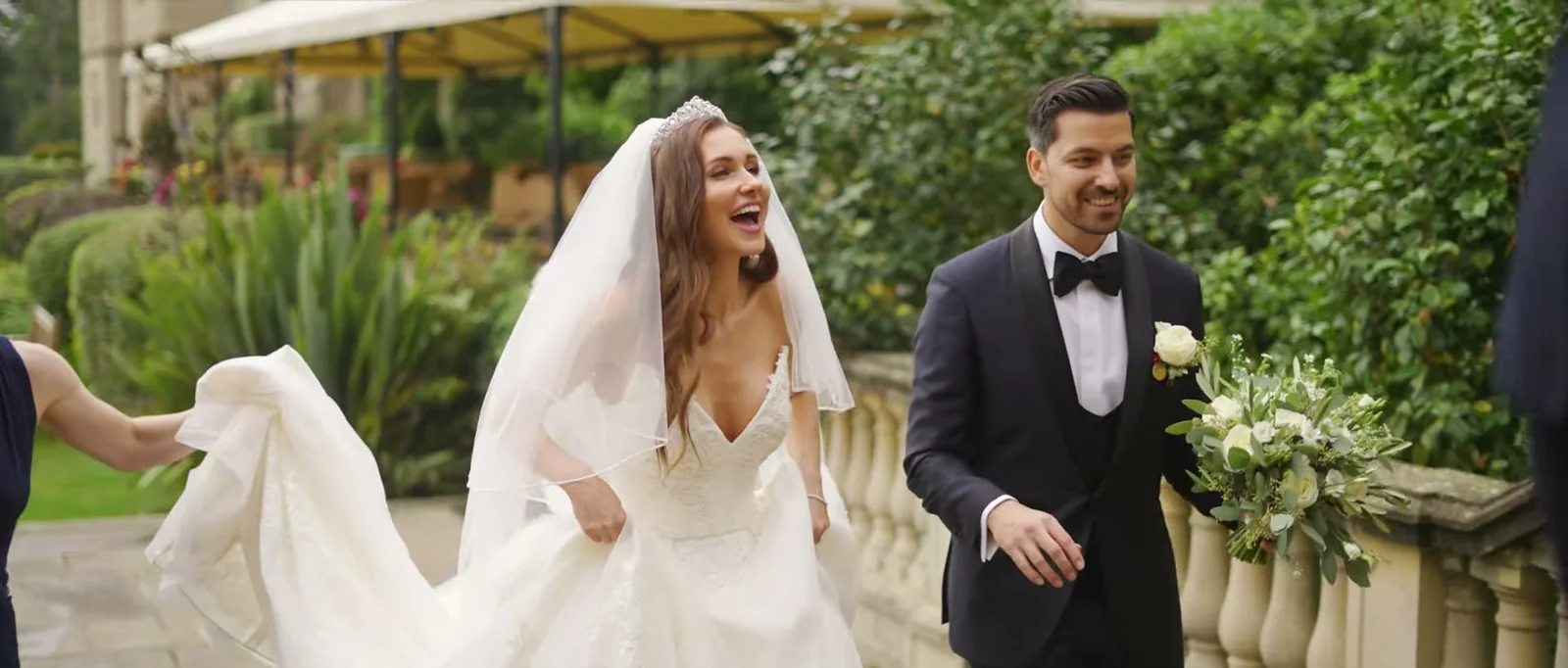 A bride laughs and holds up her long dress as she walks beside a groom along a stone balustrade on the terrace at South Lodge