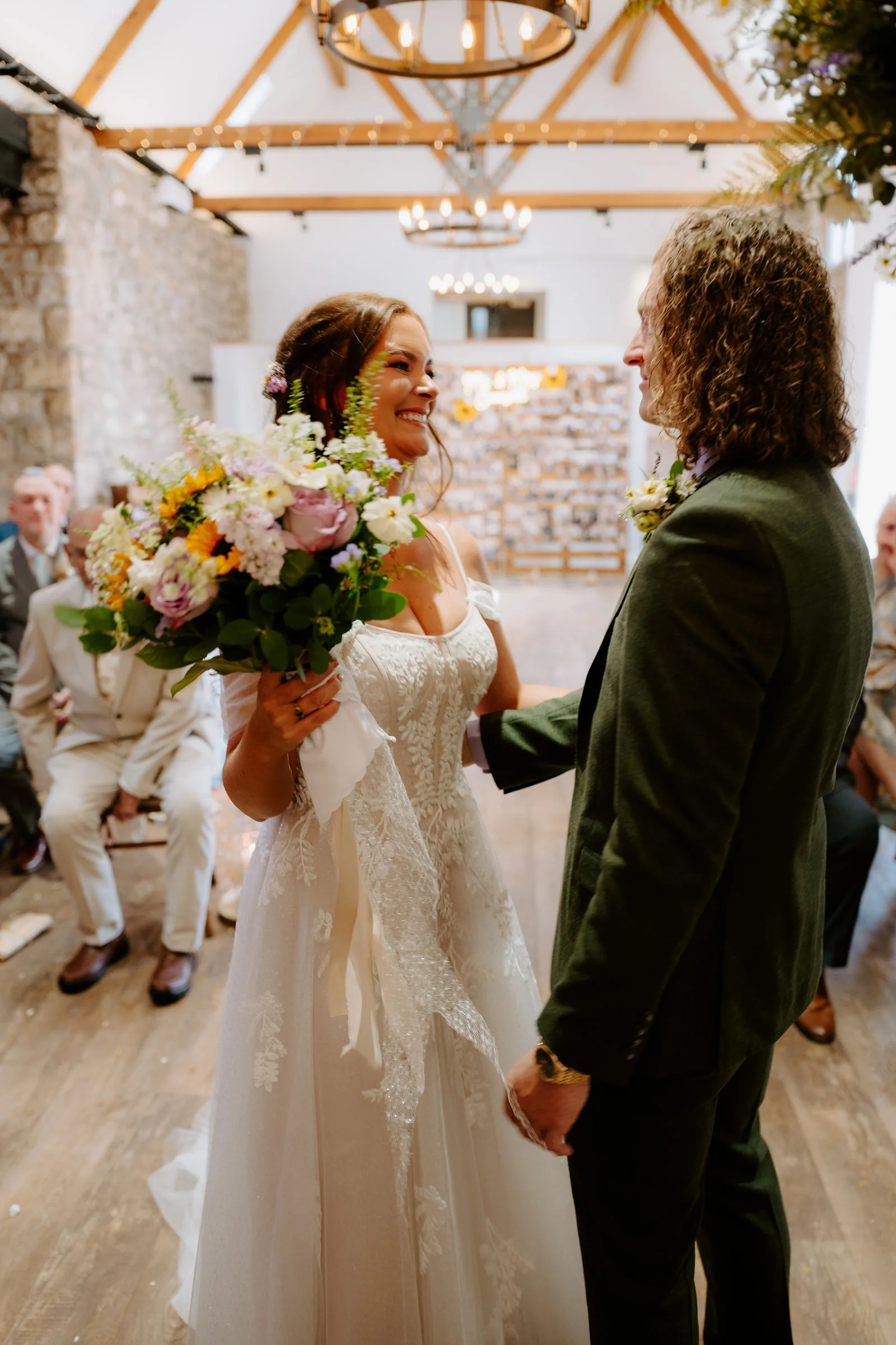 A smiling bride holding a bouquet of sunflowers and pink roses faces a groom in a dark green suit inside a stone-walled barn with timber roof trusses and iron chandeliers, guests seated behind them
