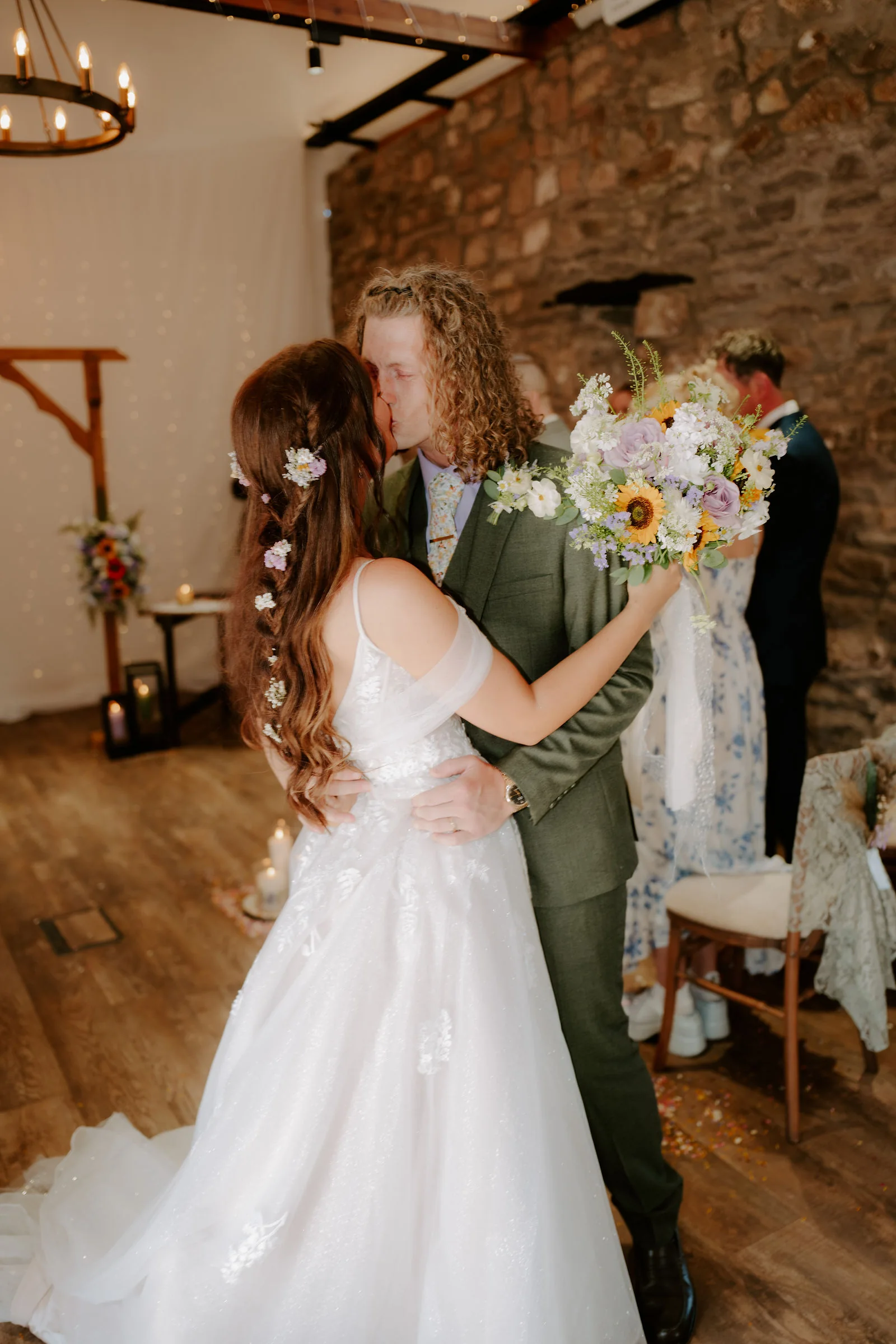 A groom in a green suit kisses his bride, who holds a bouquet of wild flowers and a sunflower, beside a stone wall and a wooden arbour dressed with flowers, under an iron chandelier