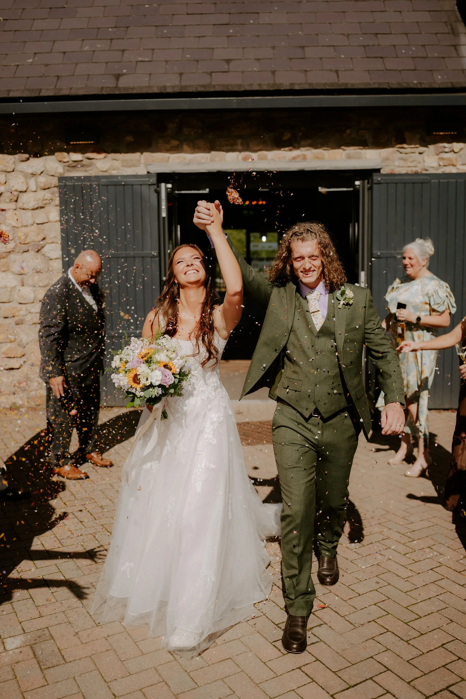 A bride in a lace gown holding a bouquet of sunflowers and pink roses and a groom in a green three-piece suit walk out of the dark double doors of a stone barn through confetti on a brick-paved yard, the bride raising her arm and a guest on each side
