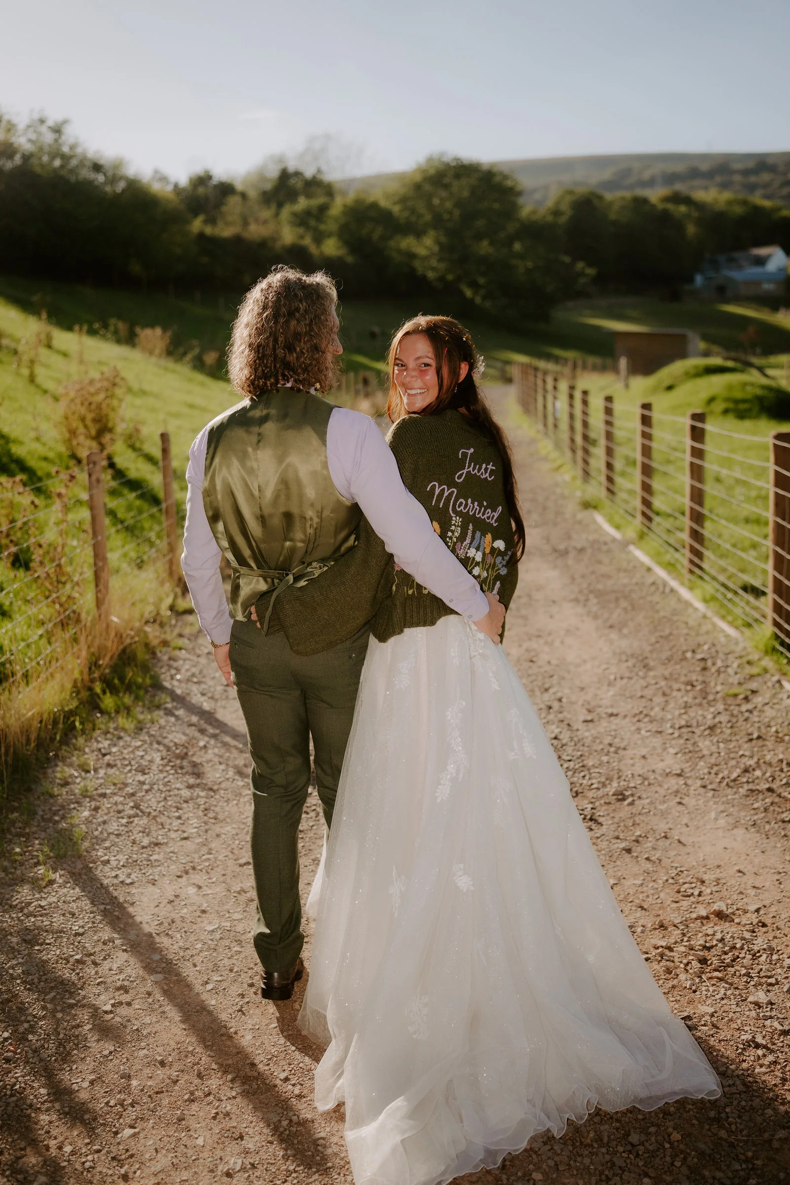 Seen from behind on a gravel track beside a wire fence in low sun, a groom in a green waistcoat has his arm around his bride, who looks back over her shoulder in a jacket lettered 'Just Married' over her tulle gown, with hills beyond
