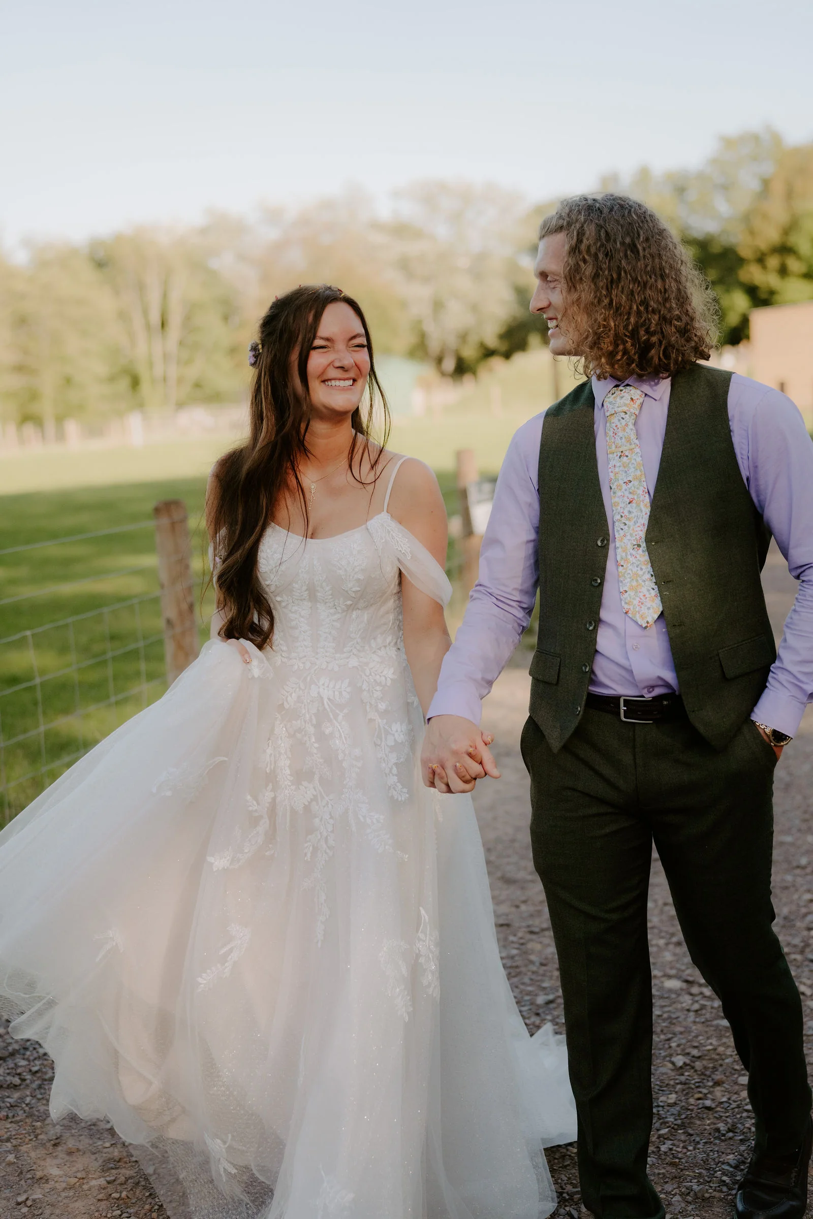 A bride in a tulle gown with off-the-shoulder straps and a groom in a green waistcoat, lilac shirt and floral tie hold hands and smile at each other on a gravel track beside a fence, with trees and a field behind