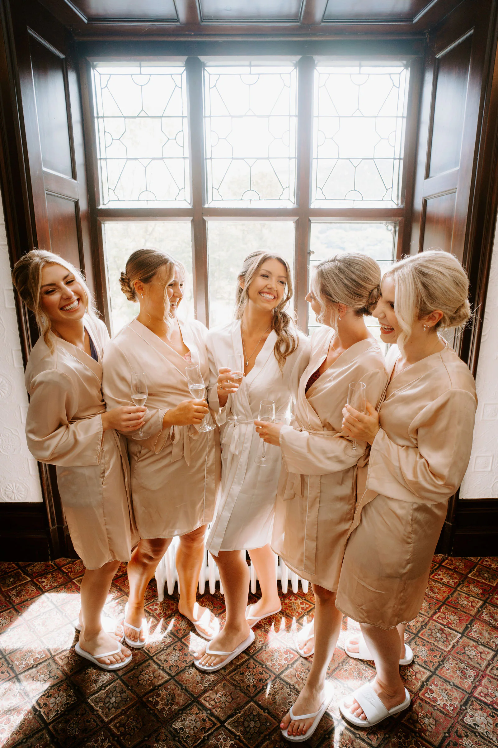 Five women in champagne satin robes laugh and hold glasses of champagne in front of a large leaded bay window, on a patterned tile floor
