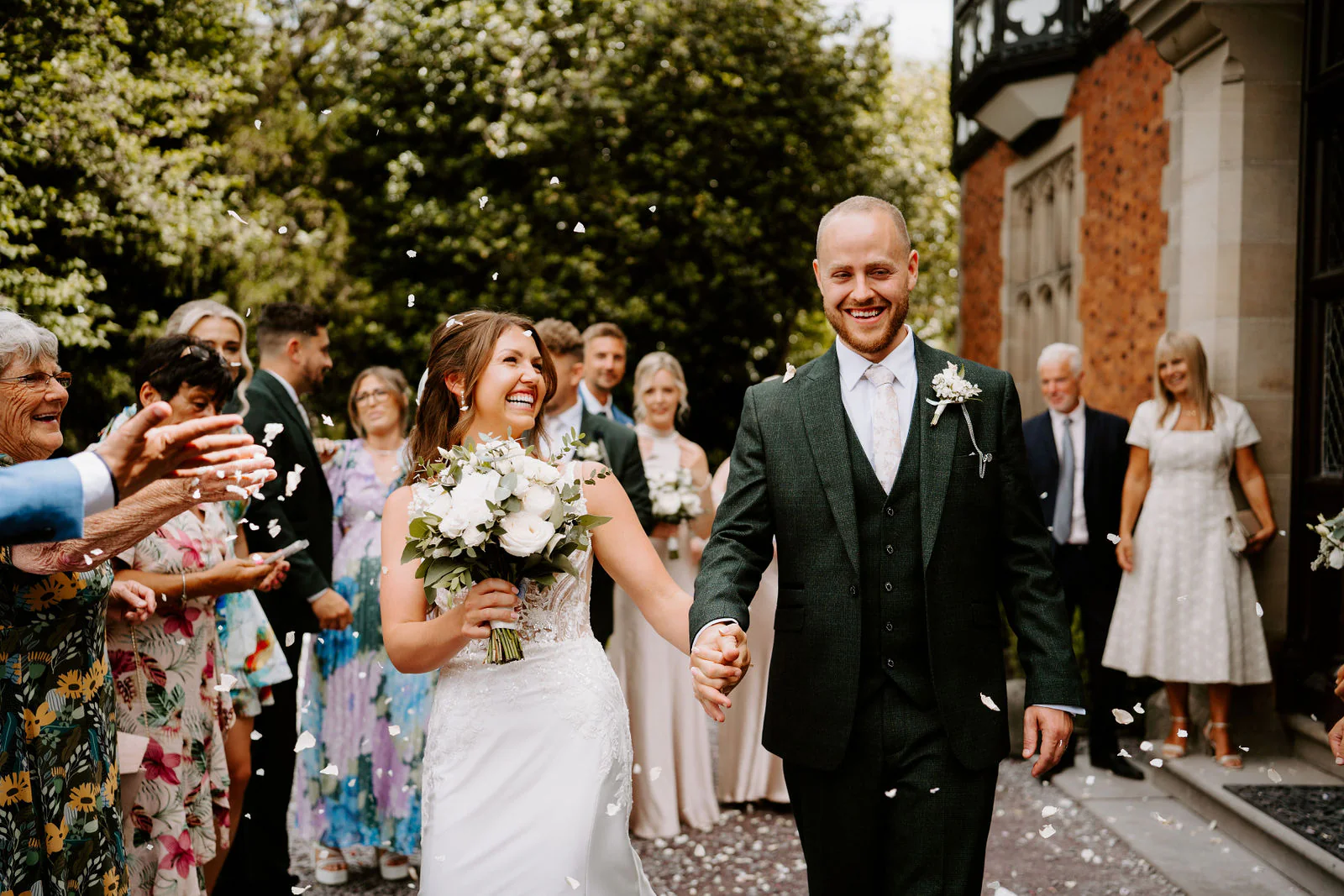 A bride and groom walk hand in hand, both laughing, through a shower of confetti between guests, with a half-timbered brick building behind them