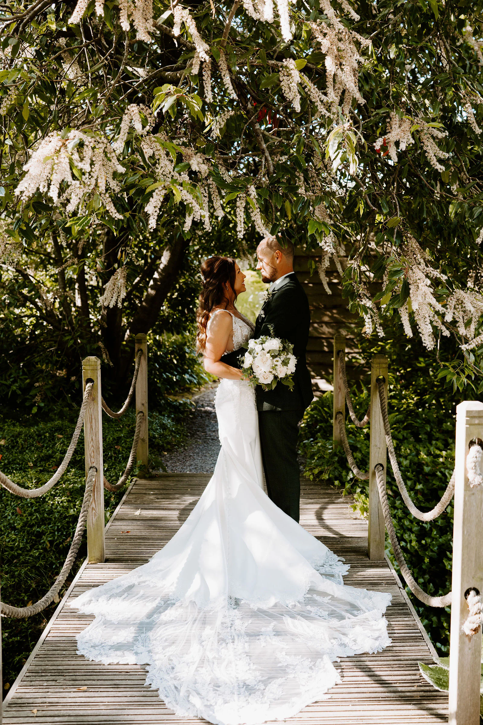 A bride in a lace gown with a very long train and a groom stand face to face on a wooden footbridge with rope handrails under a tree in white blossom