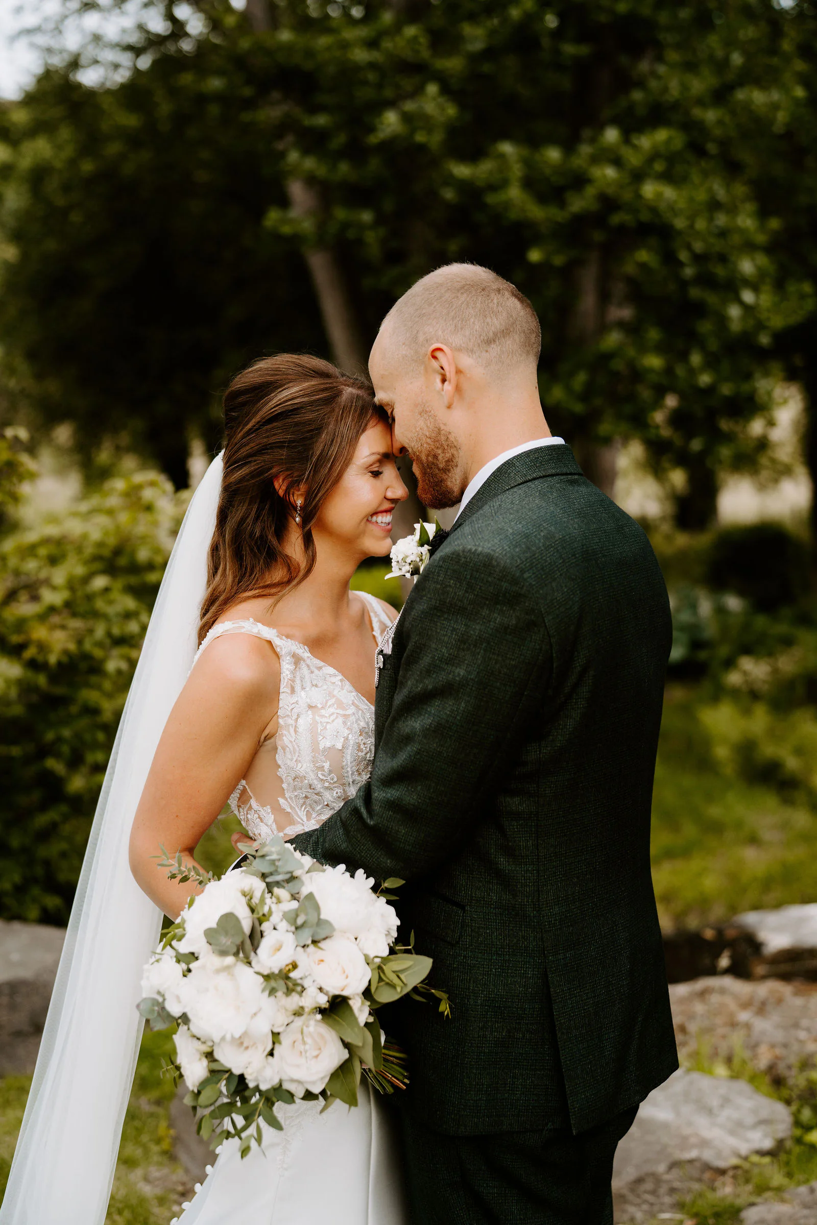 A bride in a veil and a groom in a dark green suit touch foreheads, her white bouquet in her hand, among rocks and greenery in the garden