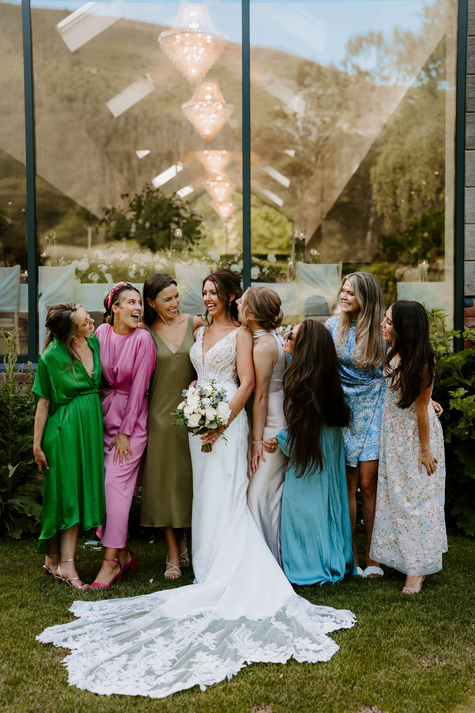 A bride with a bouquet of white flowers laughs with seven friends in colourful dresses on a lawn in front of a glass wall that reflects chandeliers and hills