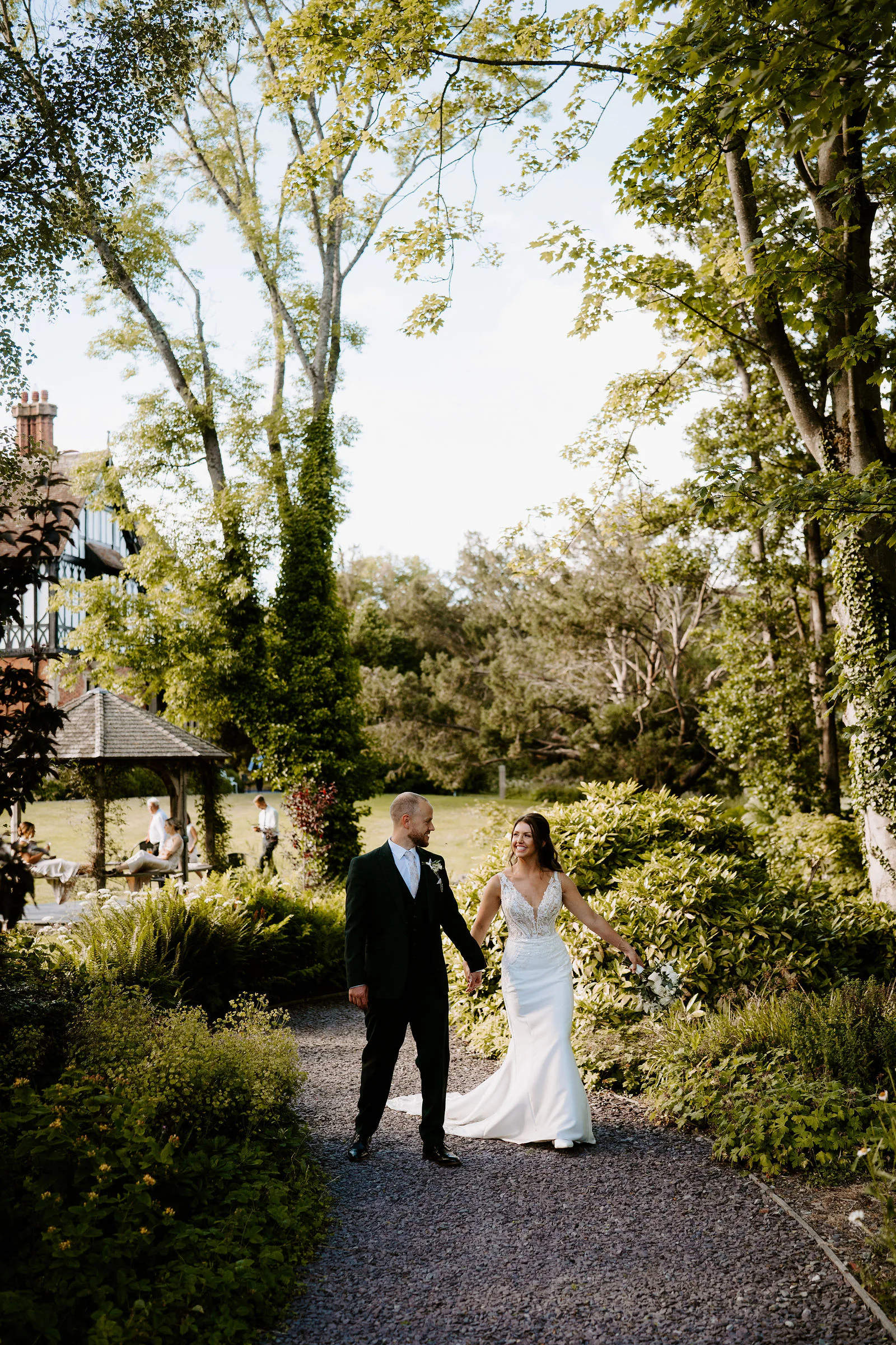 A groom in a dark suit and a bride in a white gown walk hand in hand along a gravel path in the garden, with a gazebo and a half-timbered house behind them and tall trees above