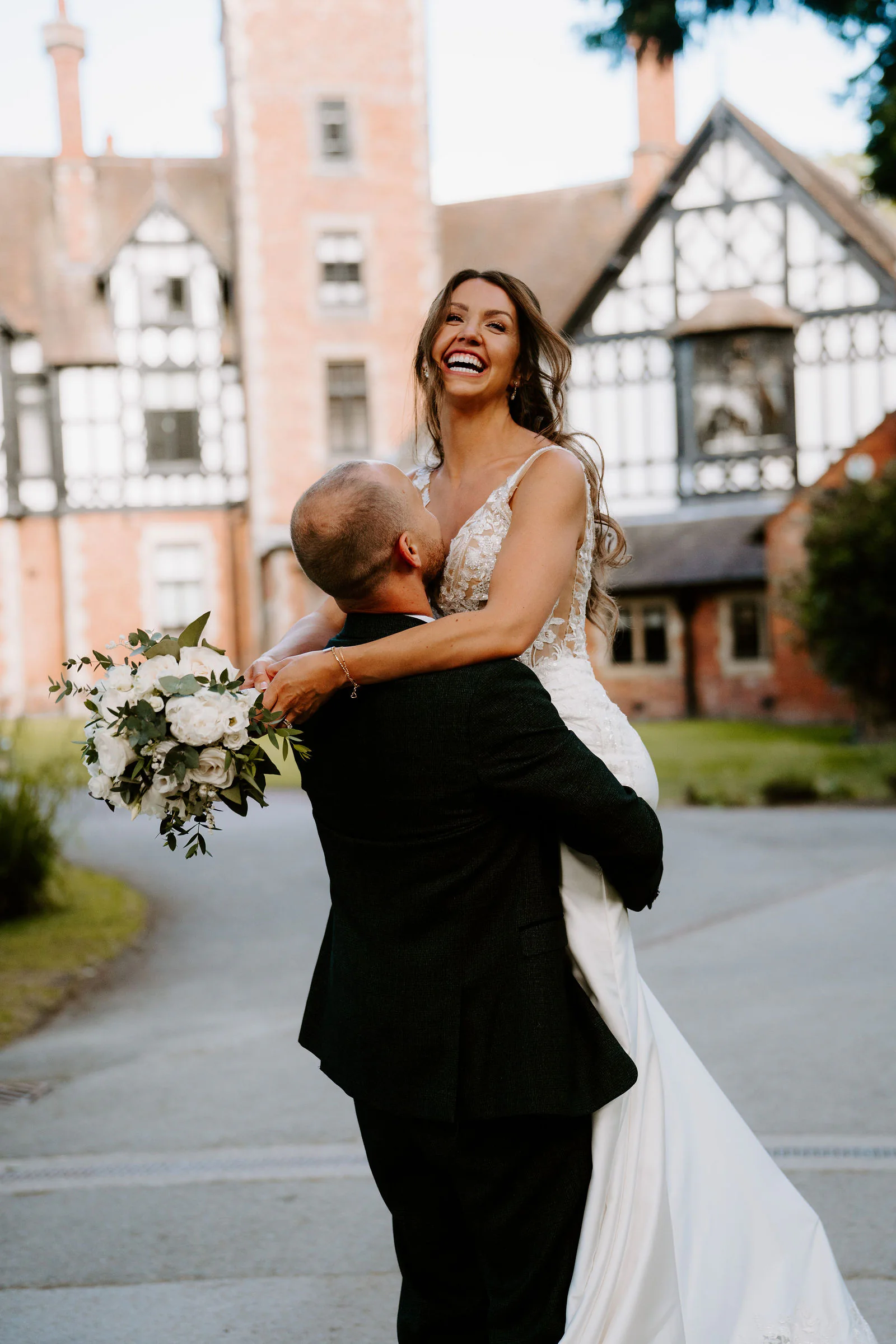 A groom in a dark suit lifts his laughing bride in his arms in front of a half-timbered brick hall